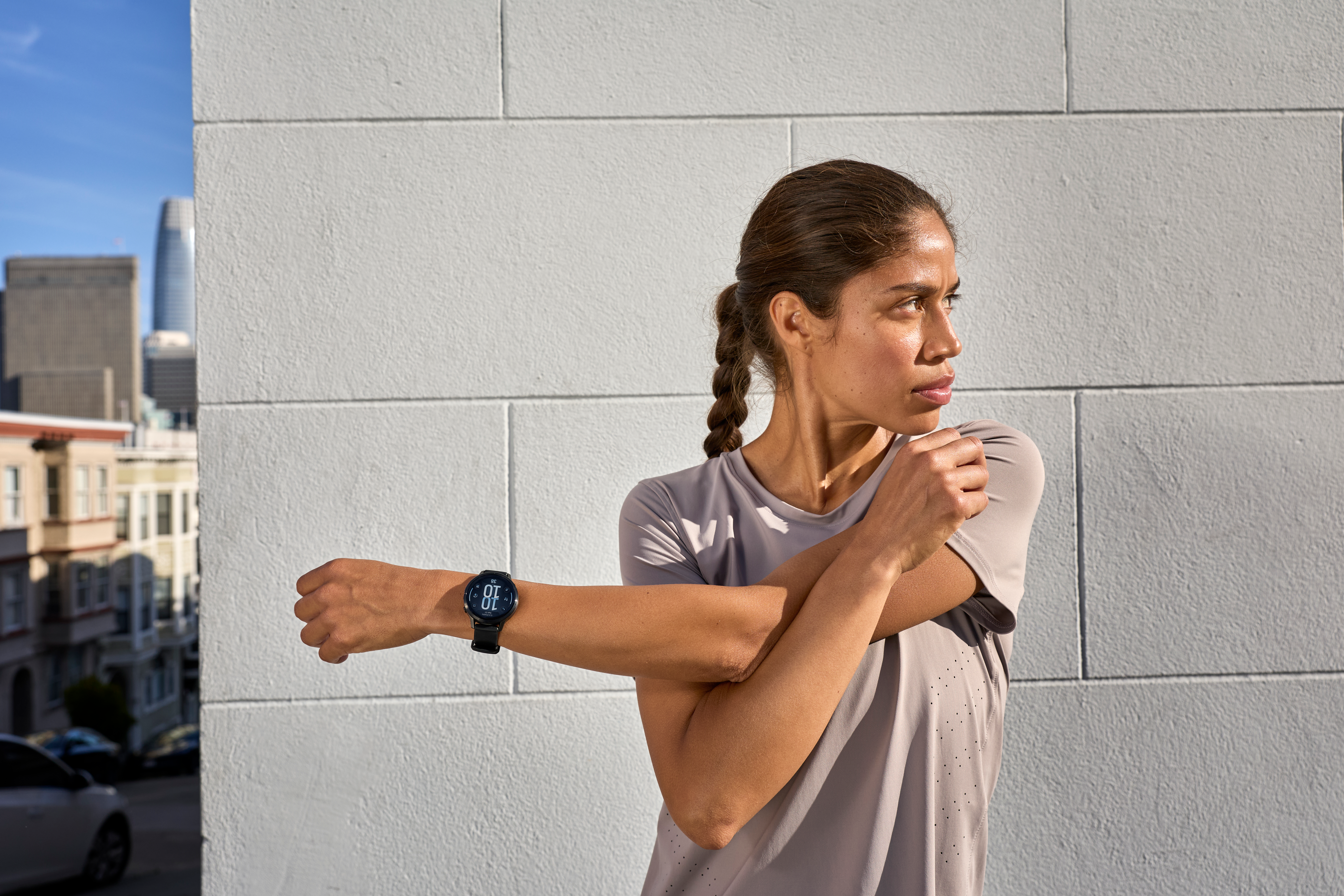 Femme s'étire le bras près d'un mur blanc. Bâtiments et ciel bleu en arrière-plan. Elle porte une montre et des vêtements de sport.