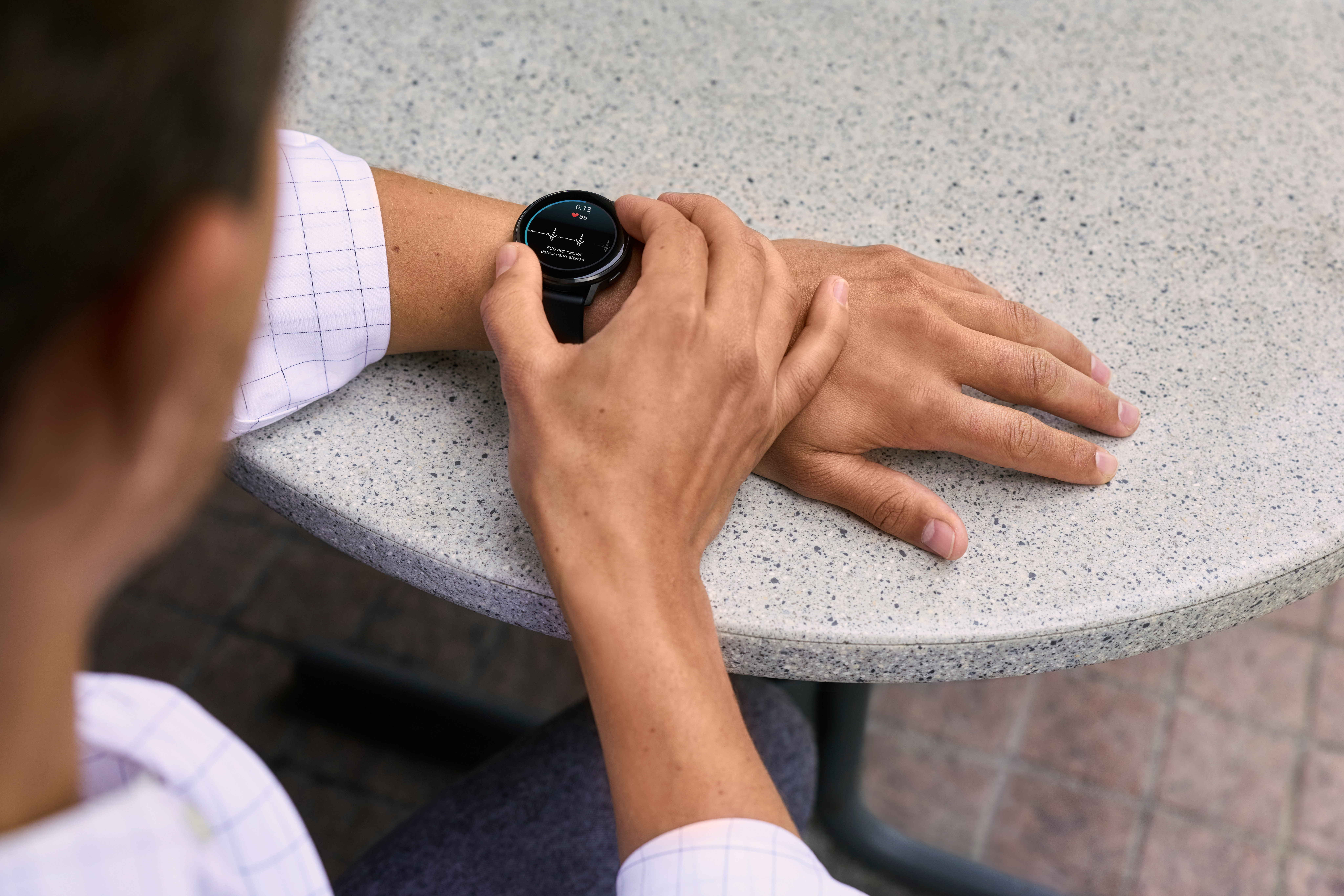 Polso di una persona con uno smartwatch che mostra parametri sanitari. La mano è appoggiata su un tavolo strutturato, con sfondo sfocato.
