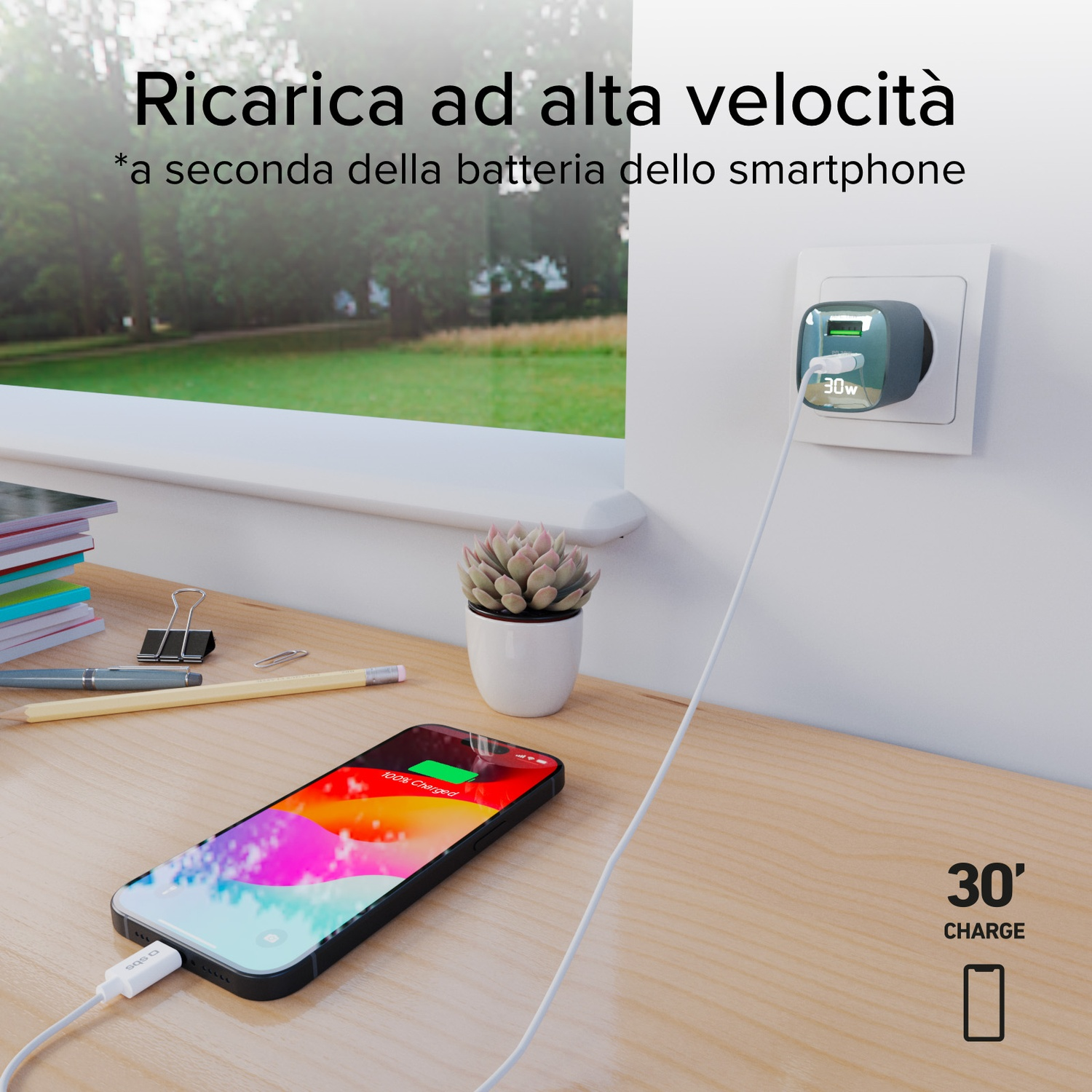 Un telefono in carica con un caricabatterie collegato alla parete. Il telefono è su una scrivania di legno con una pianta succulenta e libri.