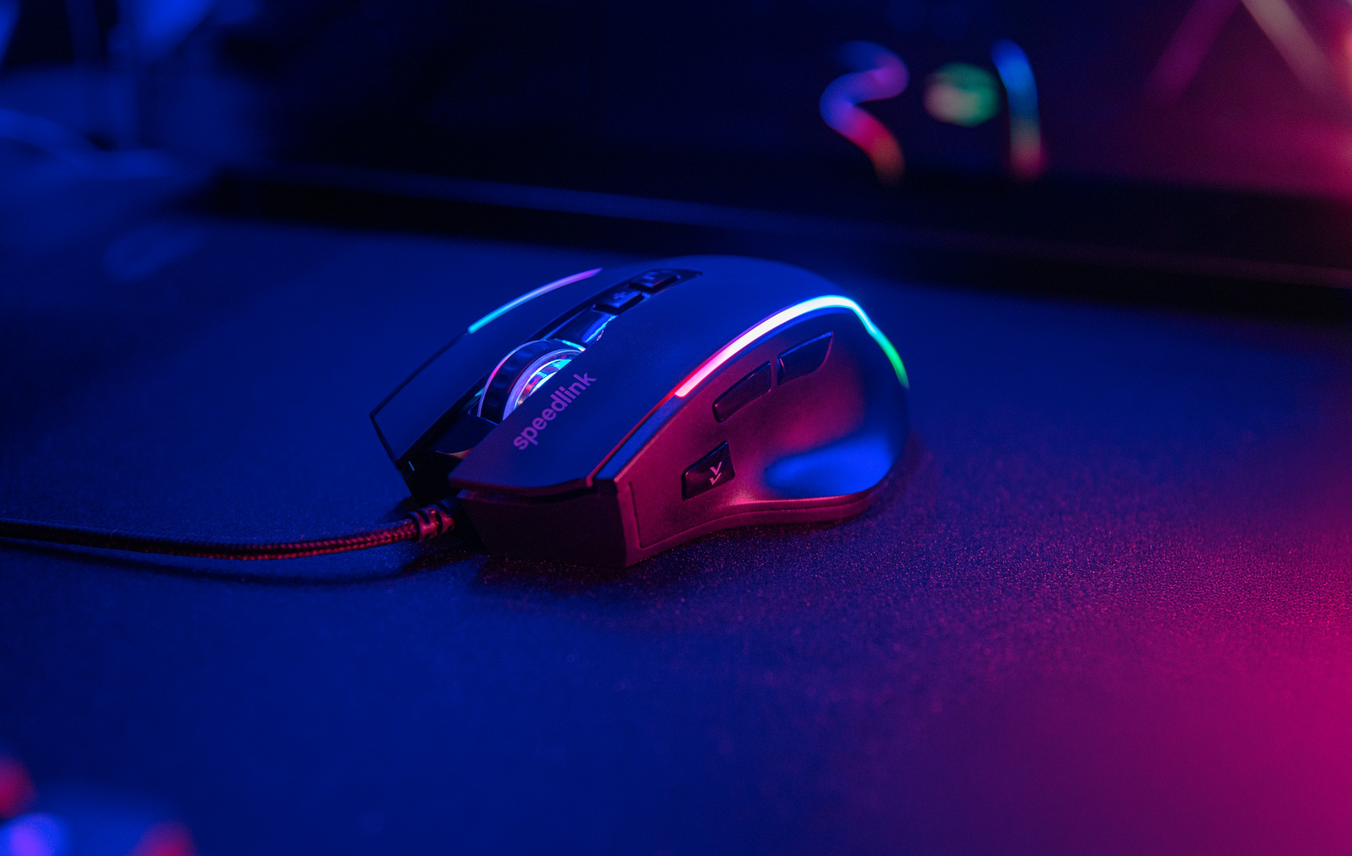 Eine schwarze Speedlink-Gaming-Maus mit RGB-Beleuchtung liegt auf einer strukturierten Oberfläche, beleuchtet von blauen und pinken Lichtern.
