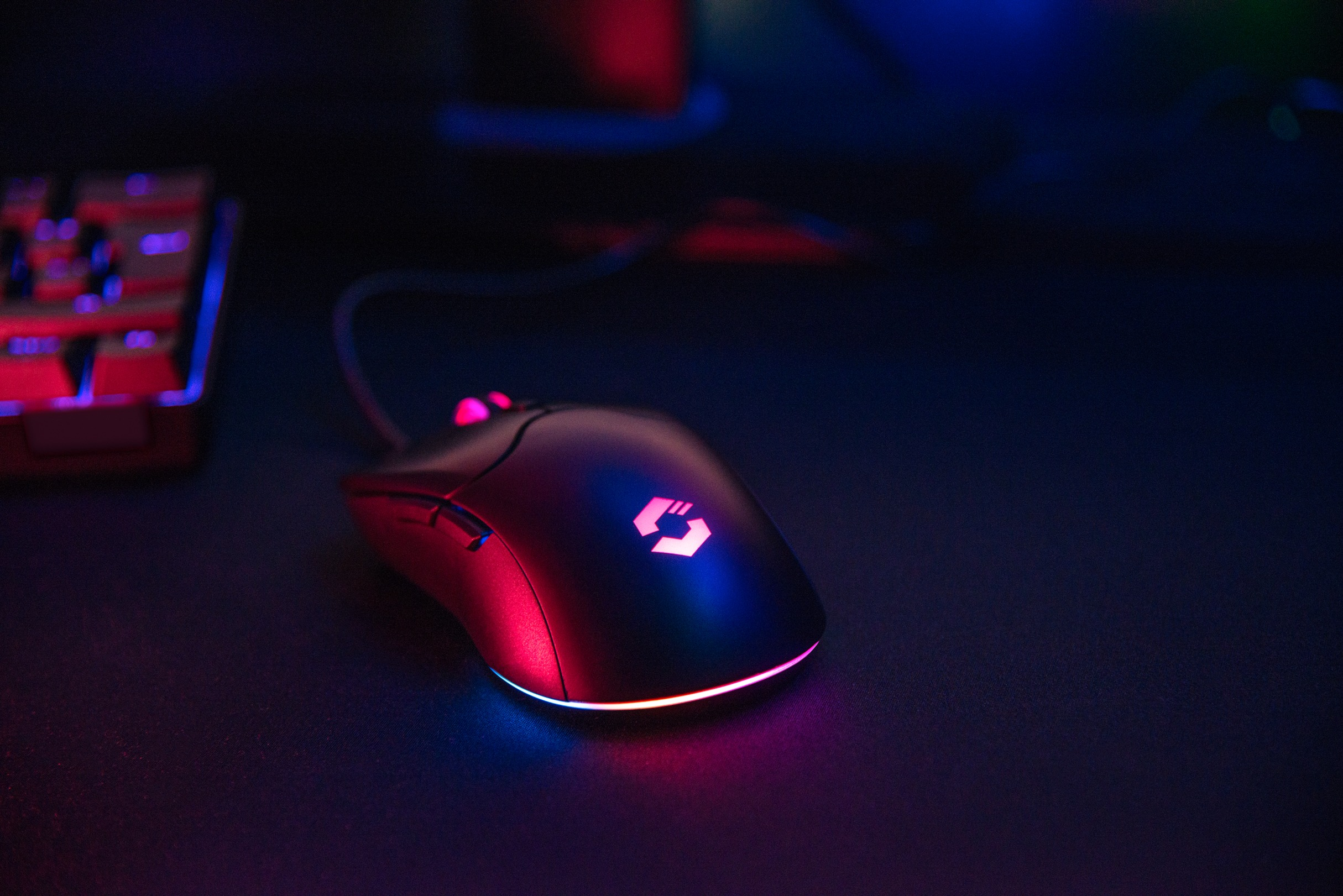 Eine Gaming-Maus mit roten und blauen Lichtern. Im Hintergrund ist eine Tastatur. Dunkle Umgebung.