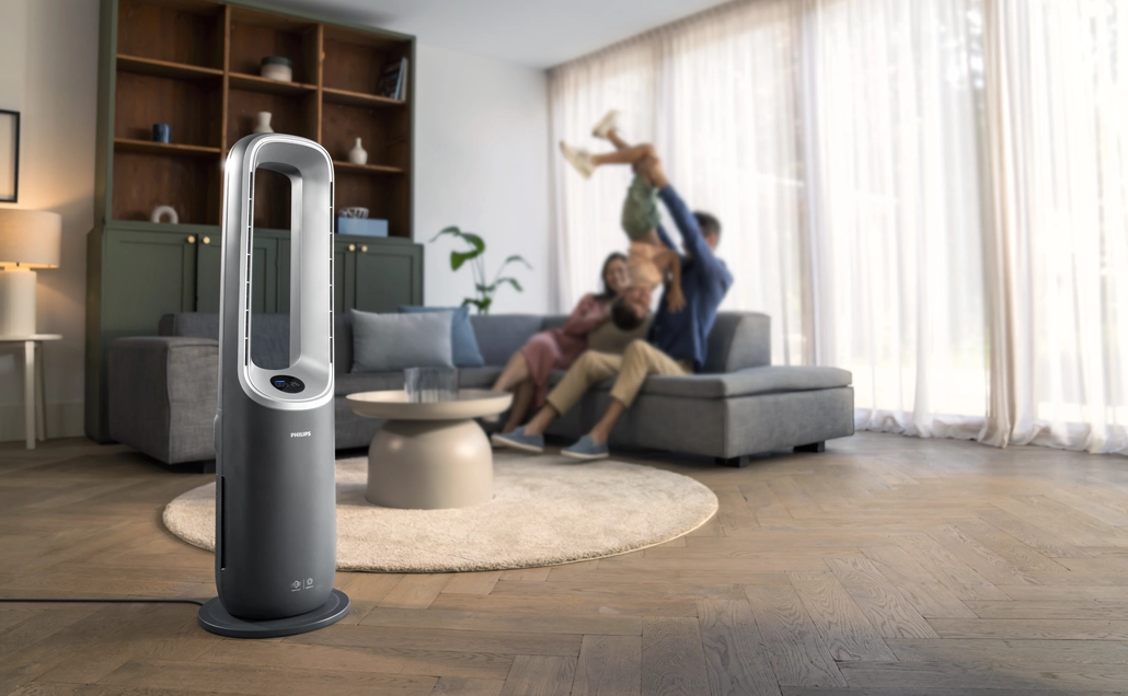 Eine Familie spielt in einem Wohnzimmer. Im Vordergrund steht ein Ventilator. Eine Familie sitzt auf einem Sofa. Ein Kind wird hochgehalten.