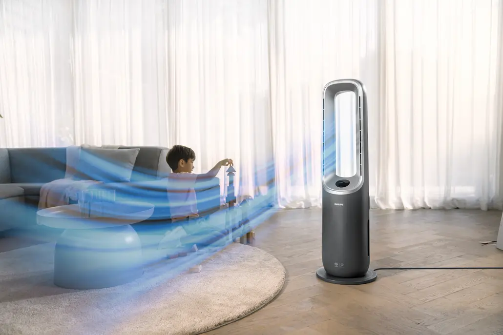 Ein Kind spielt in der Nähe eines dunkelgrauen Ventilators. Blaue Luft strömt auf das Kind zu. Im Hintergrund sind ein Sofa, ein Tisch und Vorhänge.