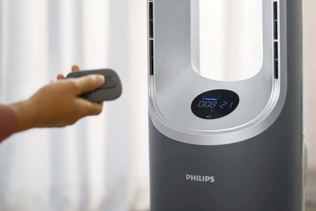 Eine Person hält eine Fernbedienung und bedient einen Philips Luftreiniger. Der Reiniger ist grau mit Digitalanzeige.