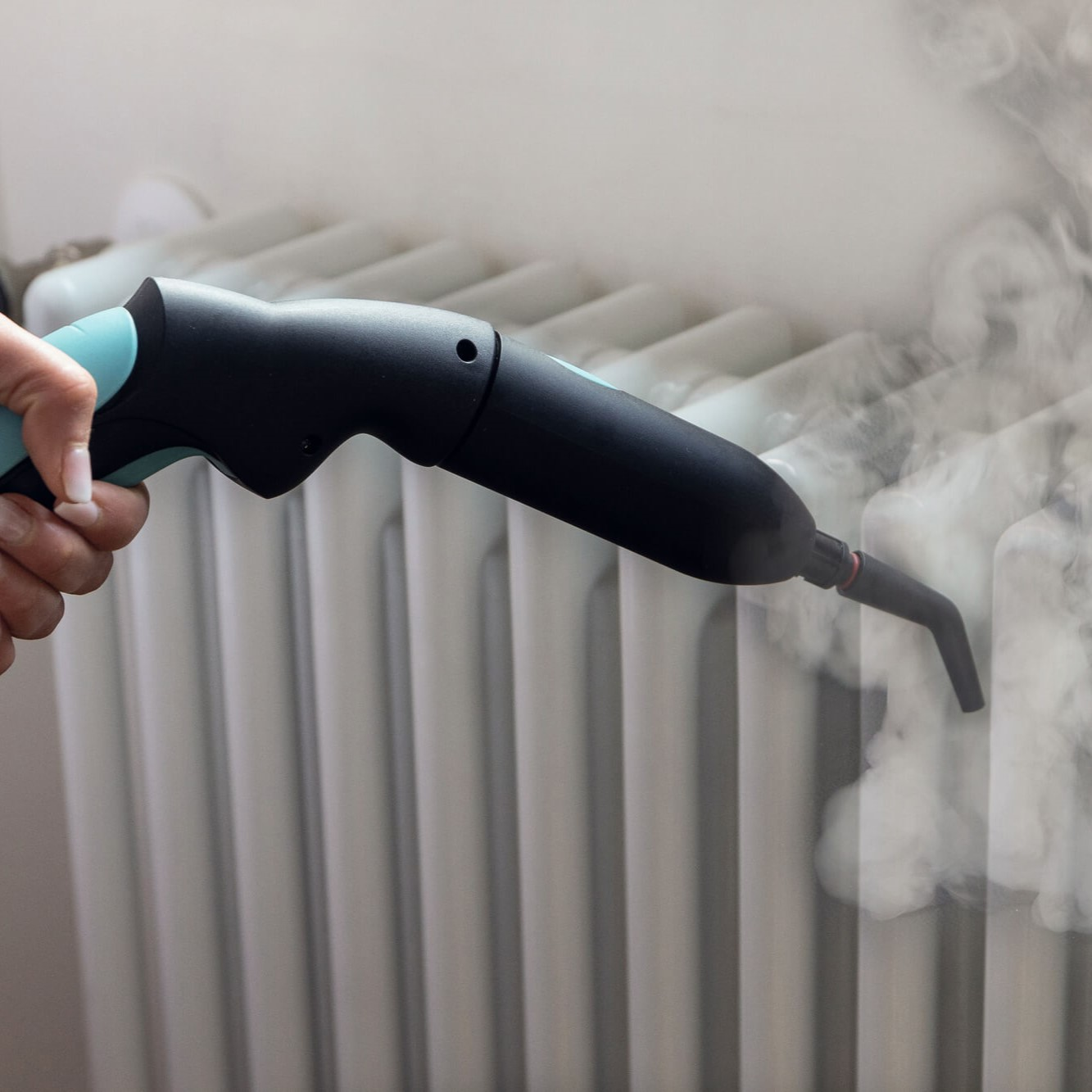 Eine Hand hält einen schwarzen und teal Dampfreiniger und gibt Dampf auf einen weißen Heizkörper ab. Der Hintergrund ist eine schlichte Wand.