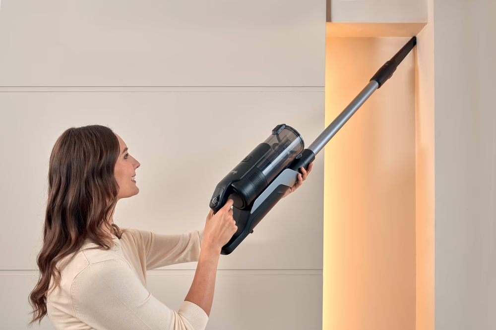Una donna usa un aspirapolvere senza fili. Sta aspirando un angolo di un muro. L'aspirapolvere è nero e grigio.