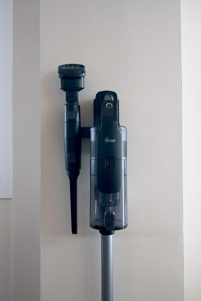 Un aspirapolvere Hoover nero e grigio è montato su una parete beige. Ha vari accessori, un contenitore per la polvere trasparente e un'asta argentata.