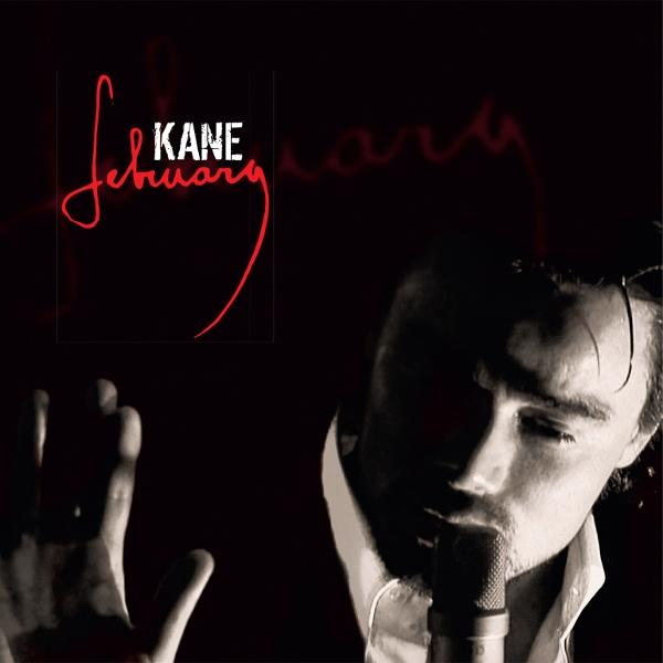 Ein Mann singt in ein Mikrofon, mit rotem Schriftzug 'February' und 'Kane'-Logo. Dunkler Hintergrund, Hand gehoben.