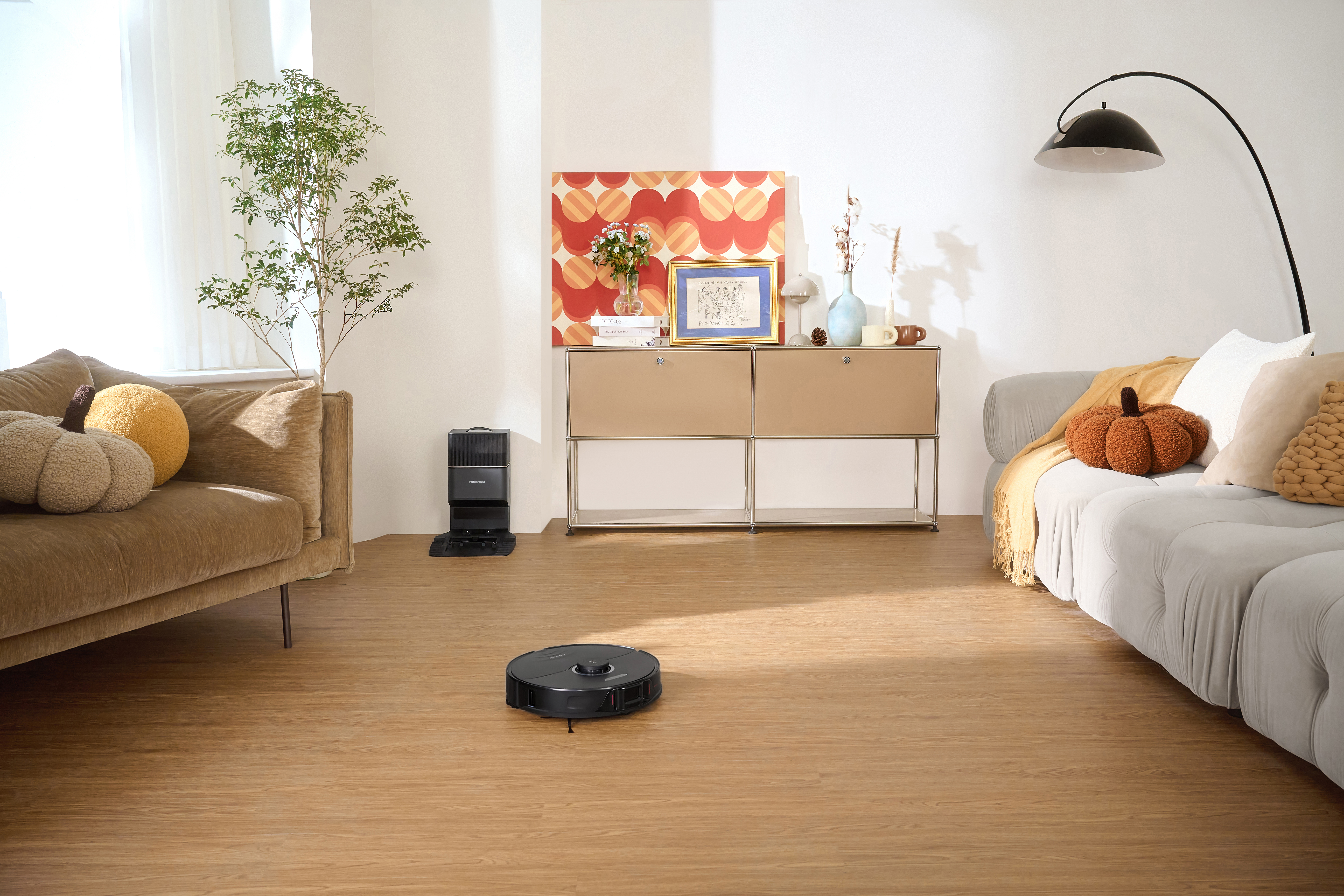 Wohnzimmer mit Staubsaugerroboter auf Holzboden. Braunes Sofa und ein Schrank mit Kunstwerk. Eine Pflanze in der Ecke.
