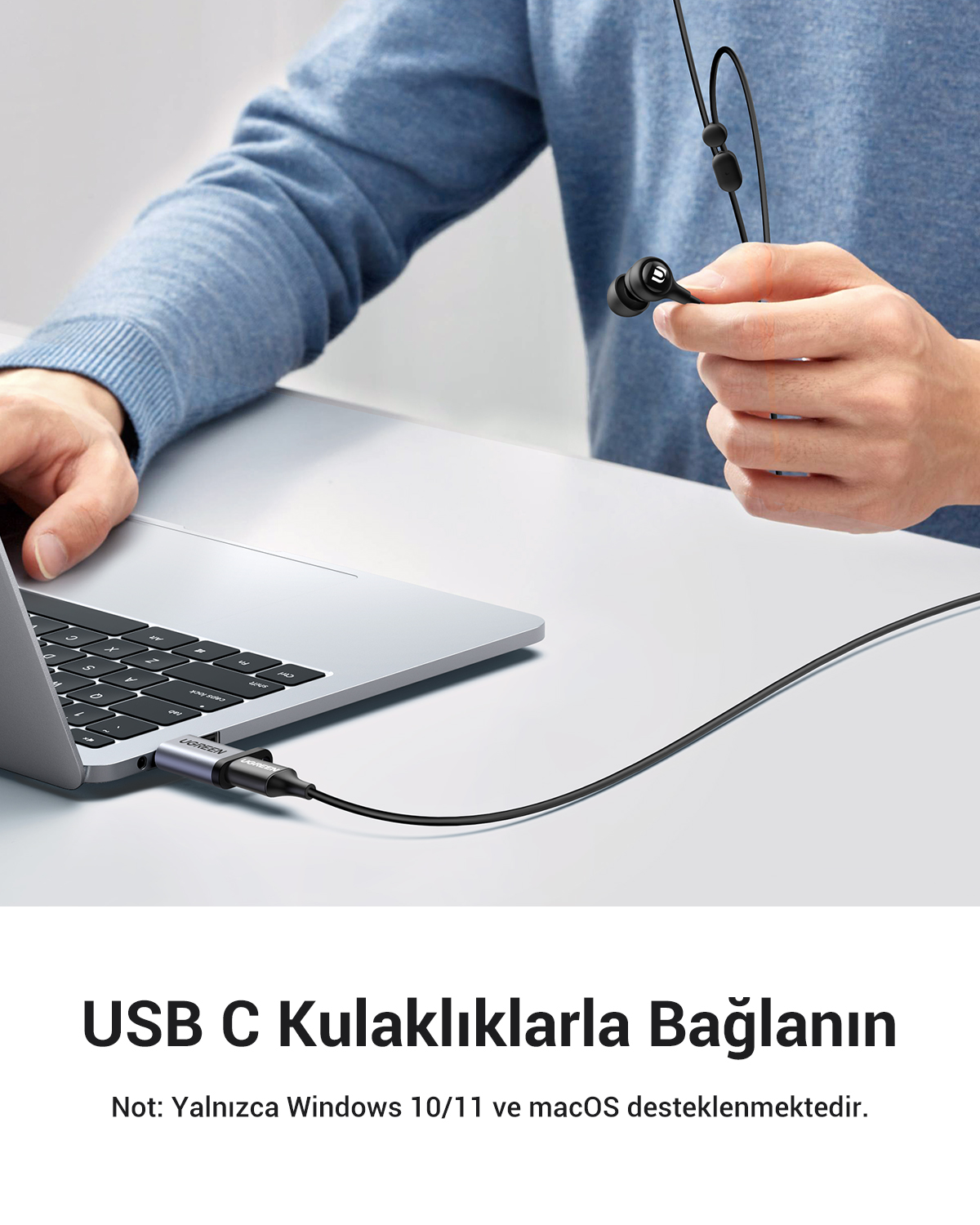 Bir kişi, USB-C kablosuyla bir dizüstü bilgisayara bağlı siyah kulaklık tutuyor. Dizüstü bilgisayar açık ve kişi mavi bir kazak giyiyor.