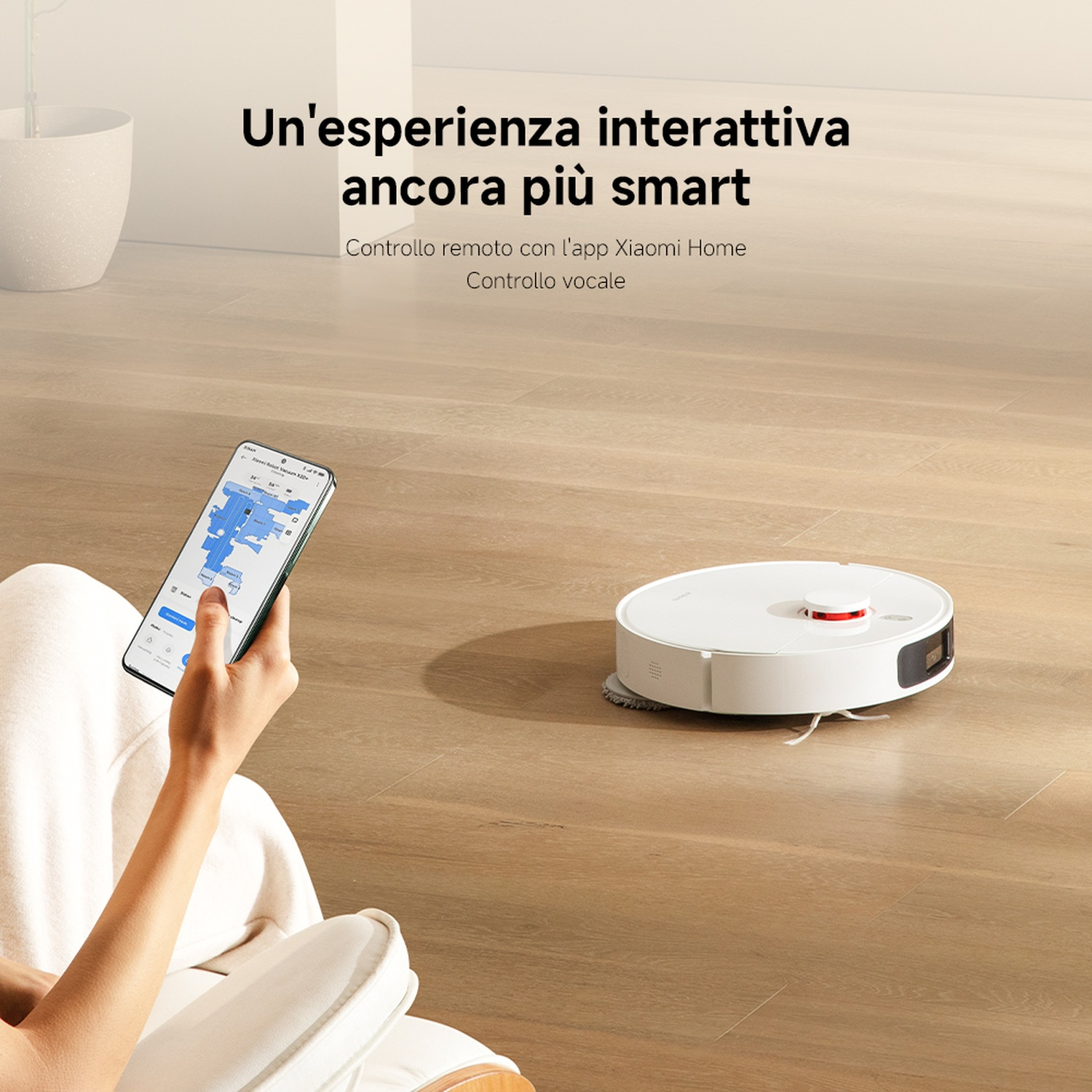 Una persona tiene in mano un telefono con un'app mappa, controllando un aspirapolvere robot bianco su un pavimento di legno.