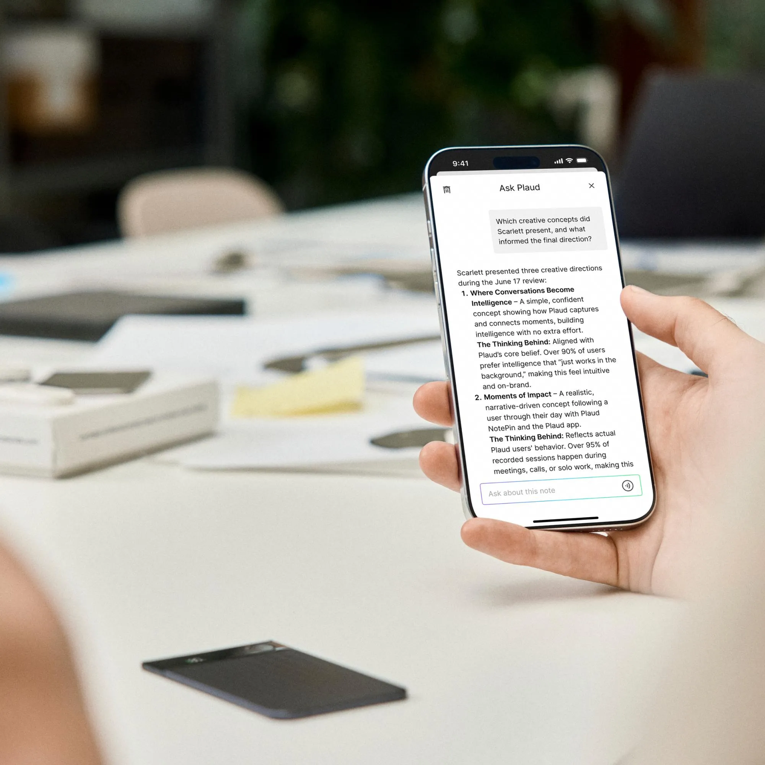 Une main tient un téléphone affichant du texte. Un appareil sombre repose sur la table. L'arrière-plan est flou.