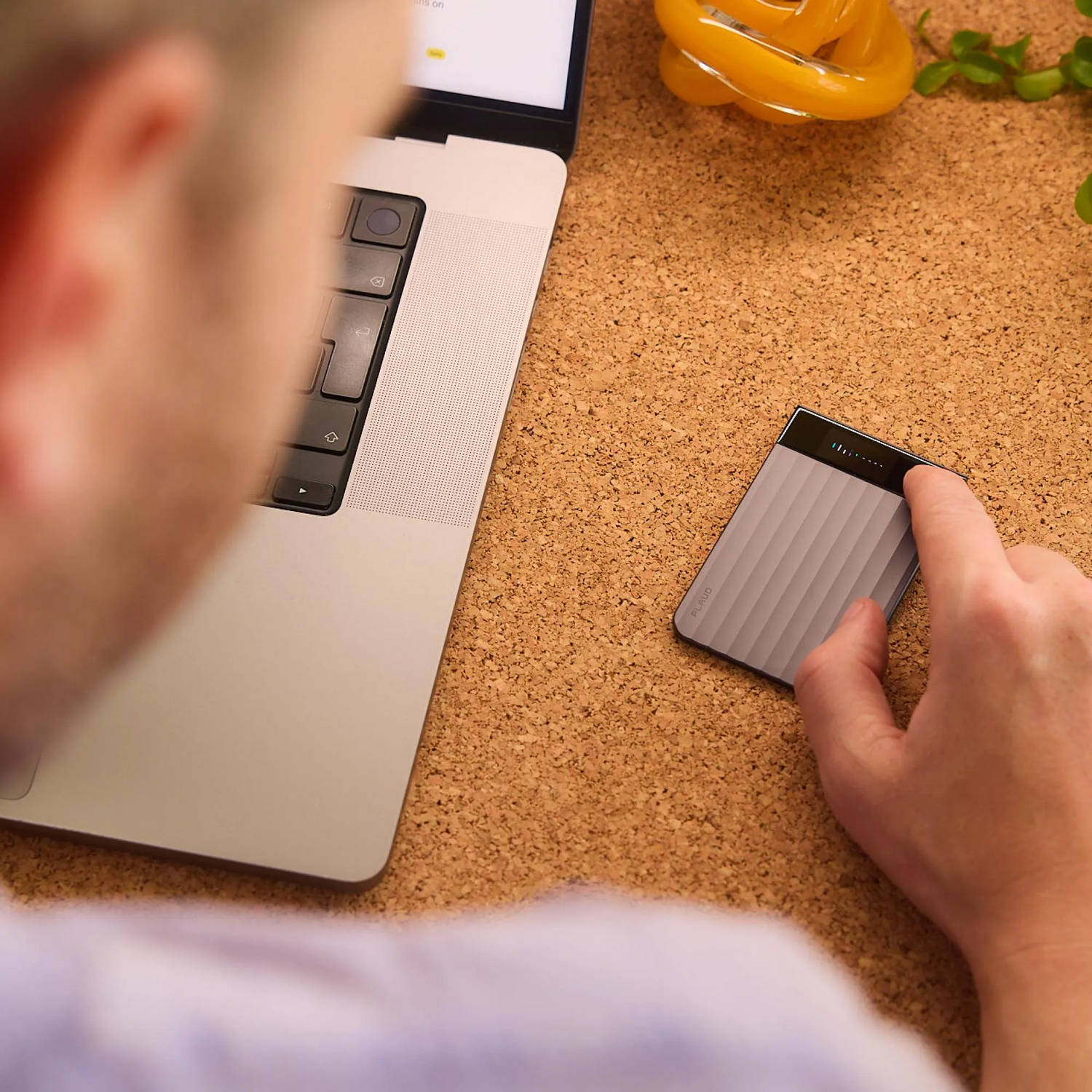 La main d'une personne touche un disque dur externe gris sur une surface en liège près d'un ordinateur portable. Un objet jaune est en arrière-plan.