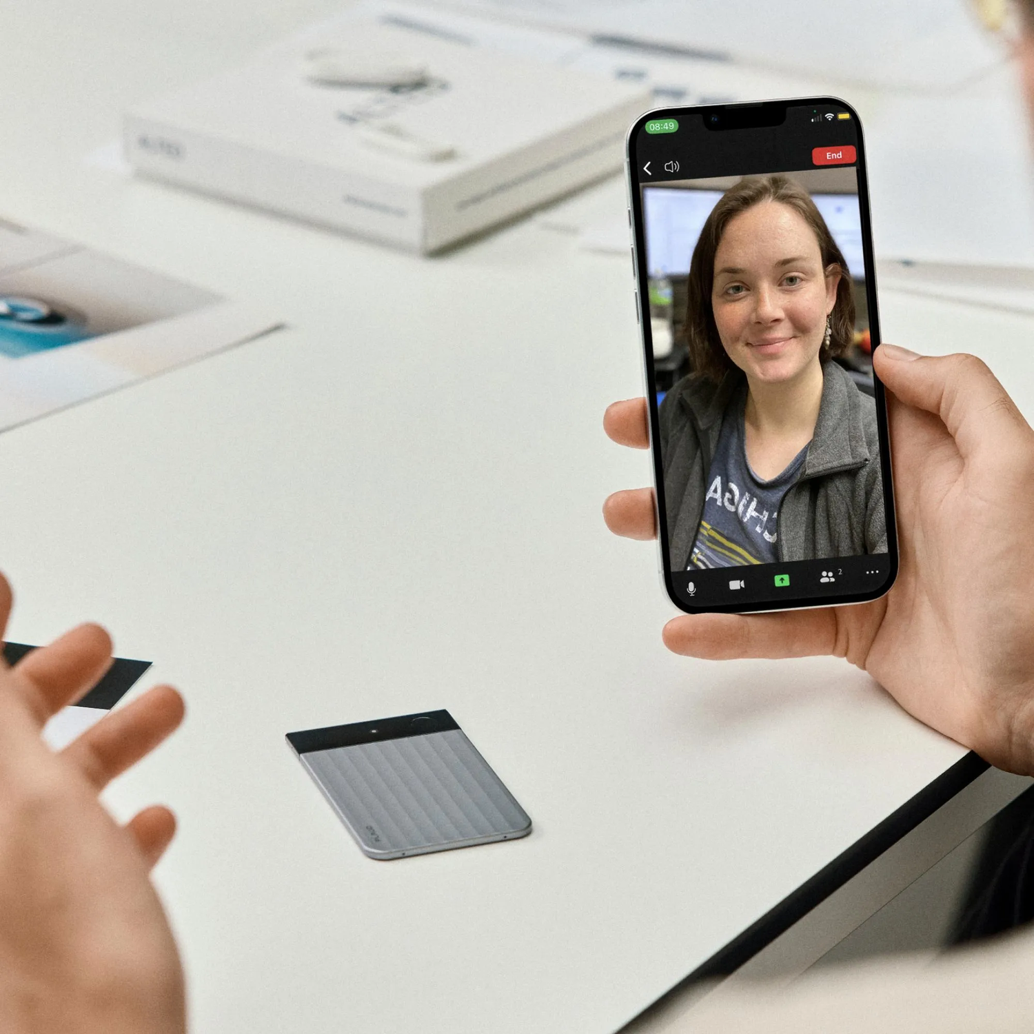 Une personne tient un téléphone affichant un appel vidéo avec une femme. Une carte grise et d'autres objets sont sur une table blanche.