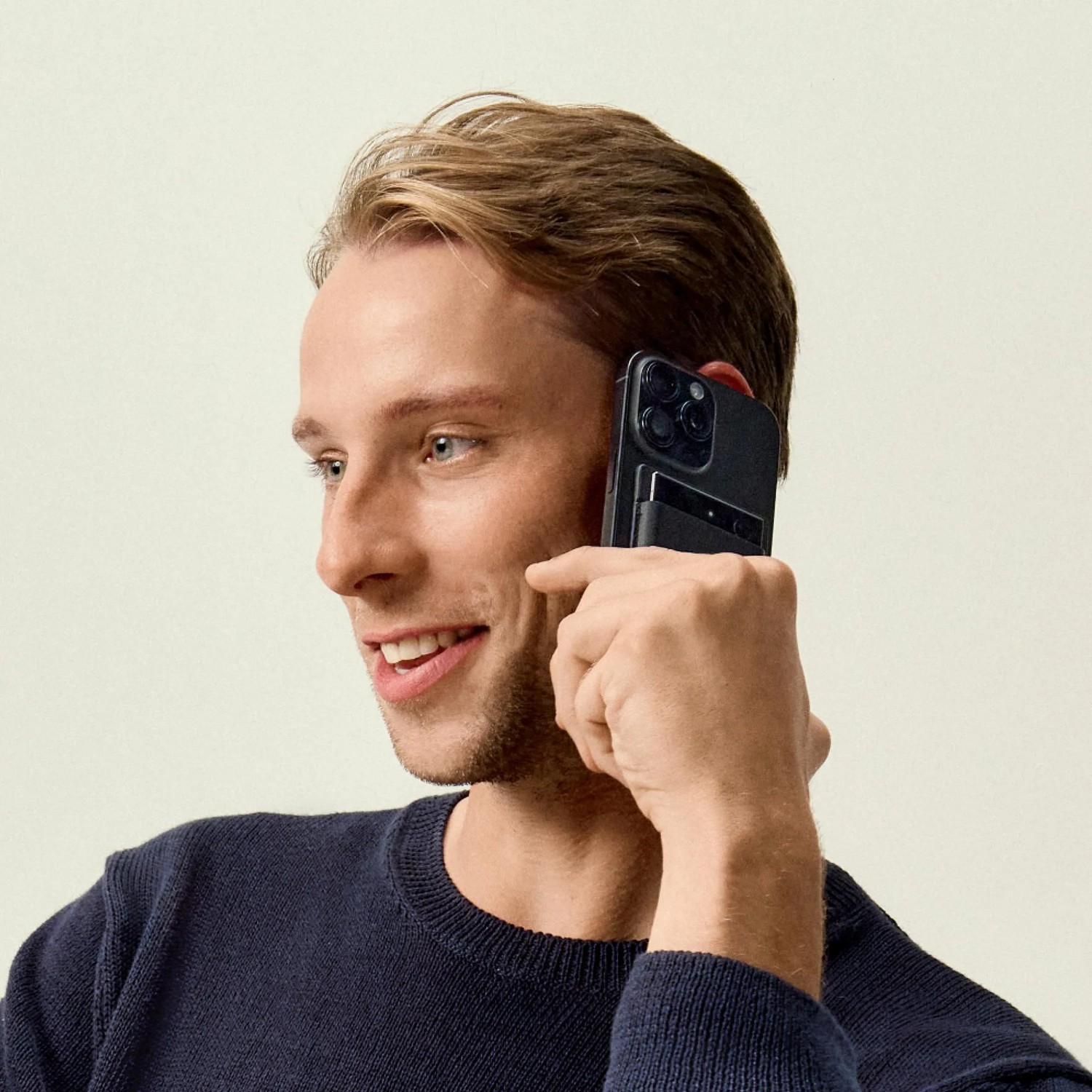 Un homme en pull bleu tient un téléphone noir à son oreille, souriant. Le fond est clair.