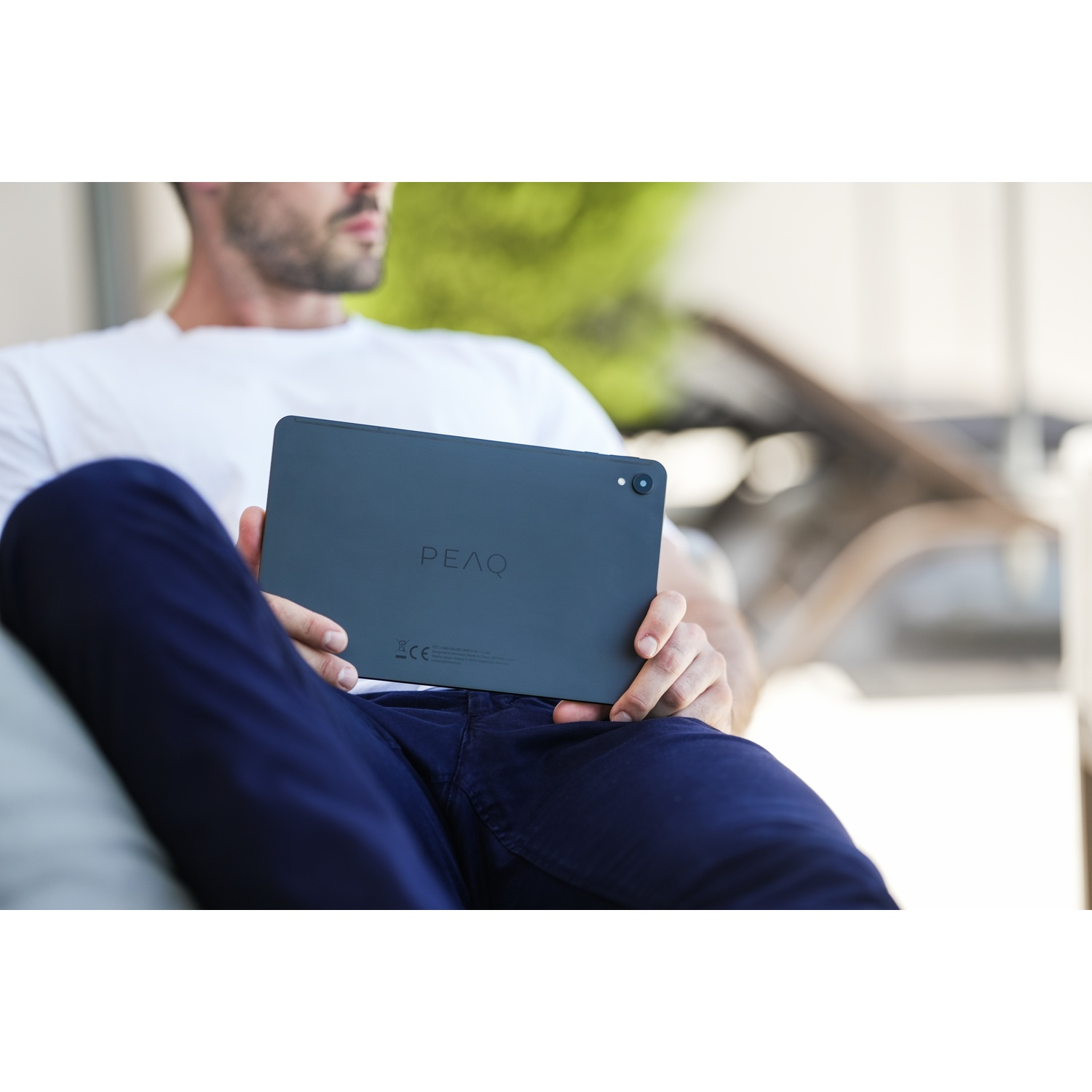 Un uomo tiene in mano un tablet scuro con la scritta PEAQ, indossa una camicia bianca e pantaloni blu, seduto fuori.