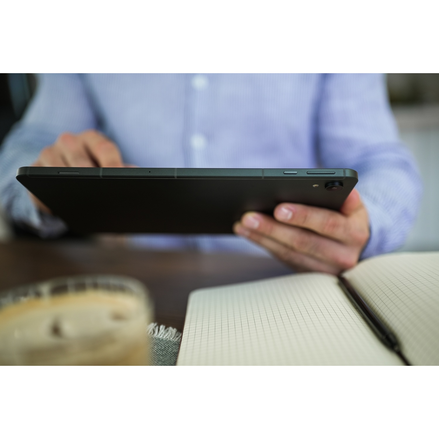 Una persona tiene in mano un tablet nero. Un blocco per appunti e una tazza di caffè sono sul tavolo. La persona indossa una camicia blu.