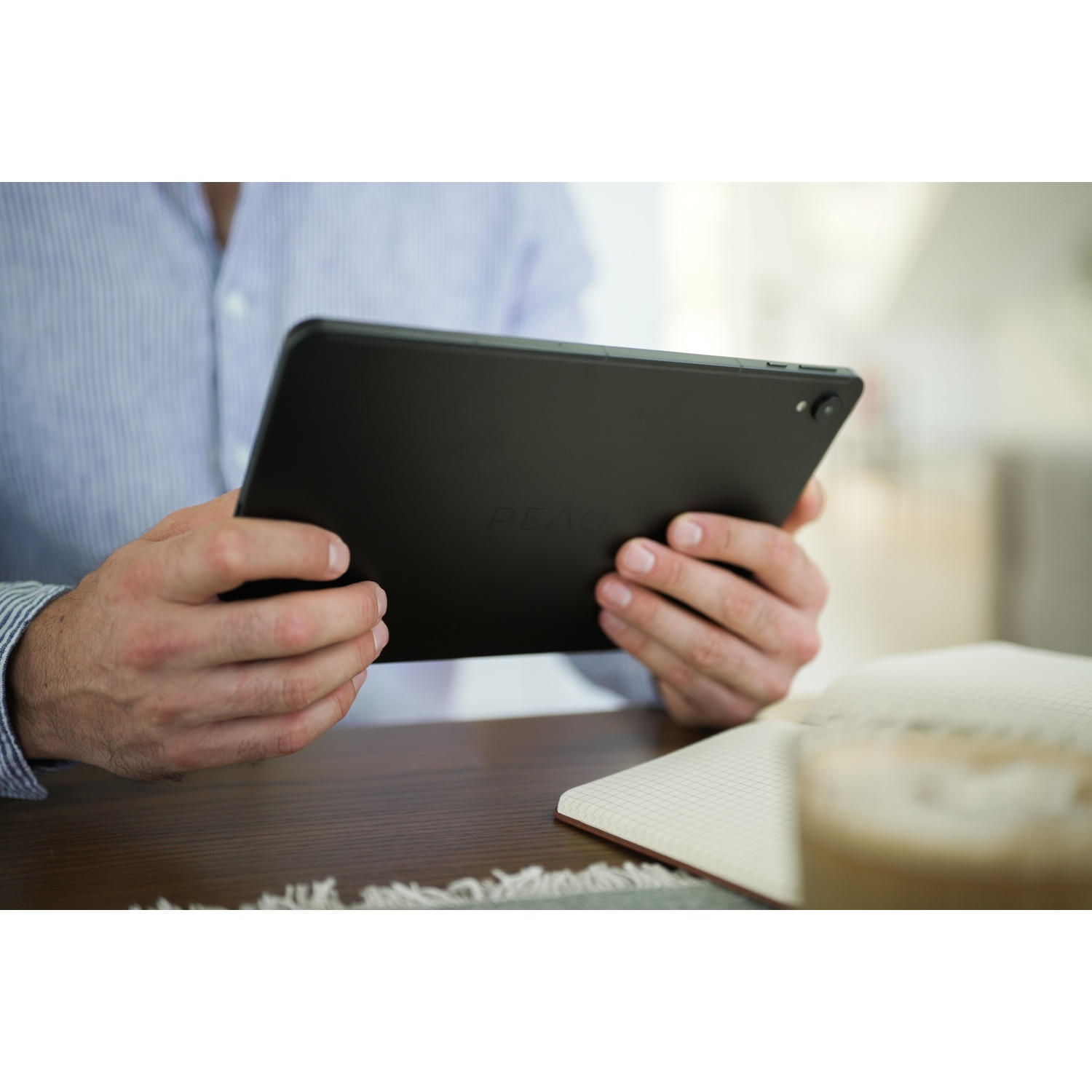 Una persona tiene in mano un tablet nero. Un quaderno e una bevanda sono sul tavolo. La persona indossa una camicia blu a righe.