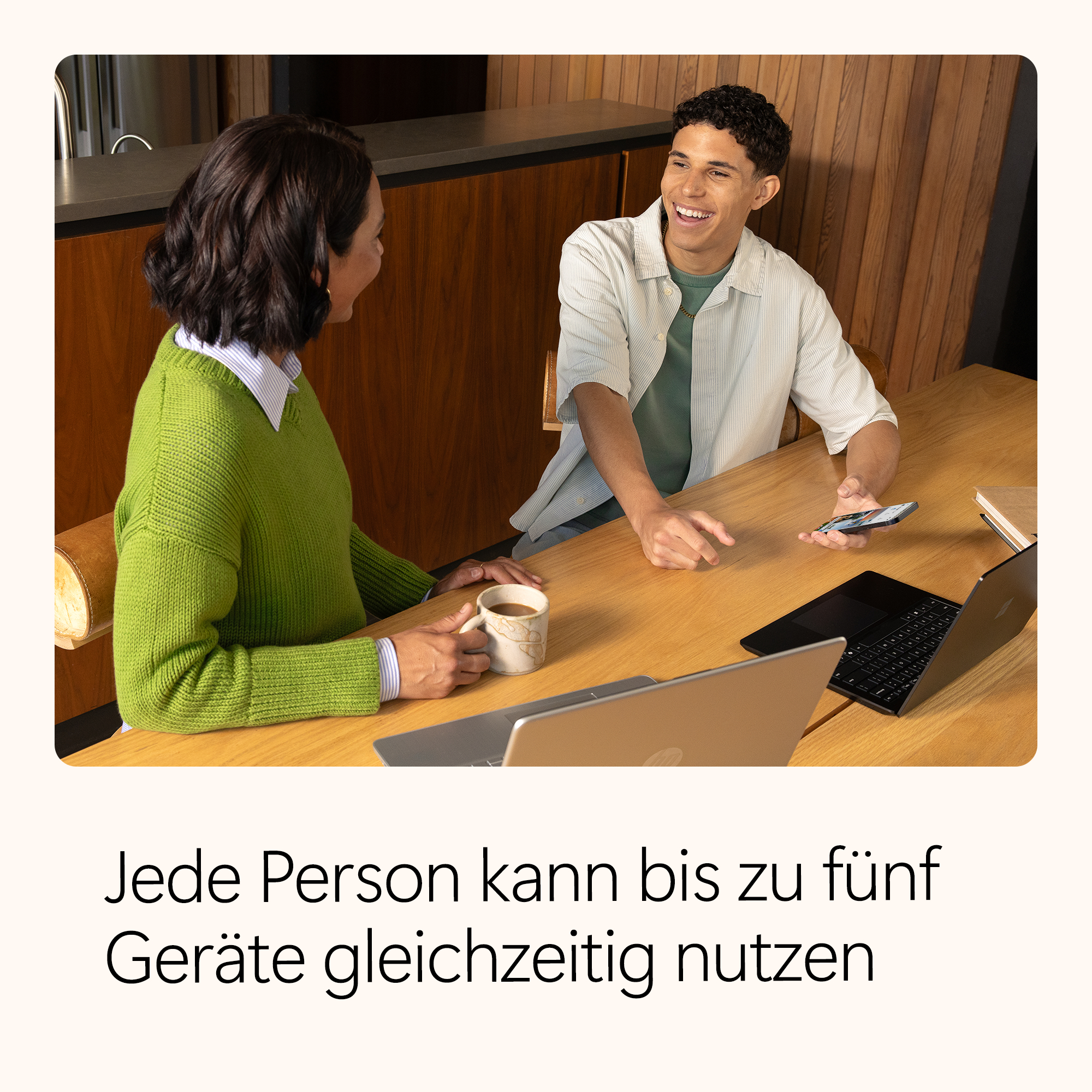 Zwei Personen an einem Tisch mit Laptops und einem Telefon. Einer hält eine Tasse, der andere lächelt. Text: Jeder kann bis zu fünf Geräte nutzen.