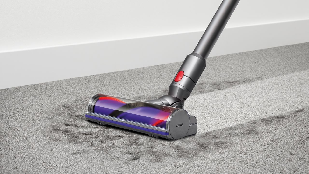 Gri bir Dyson elektrikli süpürgesi, gri bir halıyı temizliyor. Elektrikli süpürgenin başlığında kırmızı ve mor bir rulo ve gri bir sap var.
