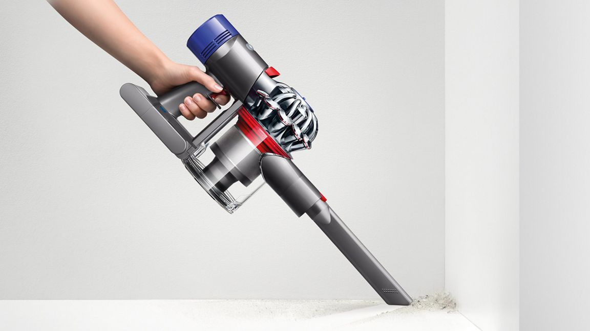El bir Dyson elektrikli süpürge tutuyor. Gri, mavi ve kırmızı. Elektrikli süpürge, beyaz bir duvarın köşesinden kiri temizliyor.