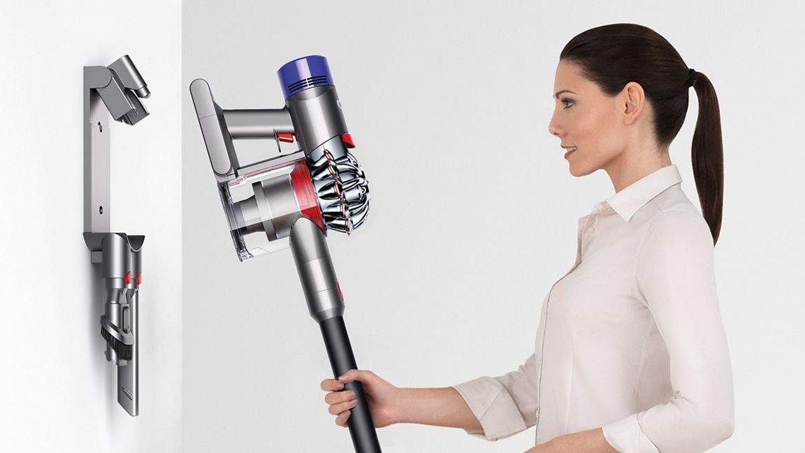 Bir kadın Dyson elektrikli süpürge tutuyor. Solda bir duvar montajı var. Elektrikli süpürge gümüş, kırmızı ve mavidir.