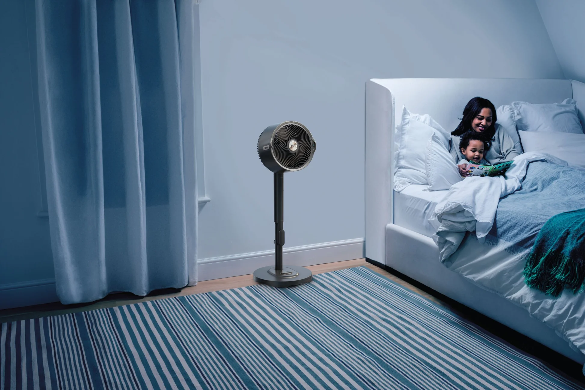Hálószoba jelenet. Egy nő és egy gyermek olvas az ágyban. Egy ventilátor áll a közelben. Kék-fehér csíkos szőnyeg a padlón.