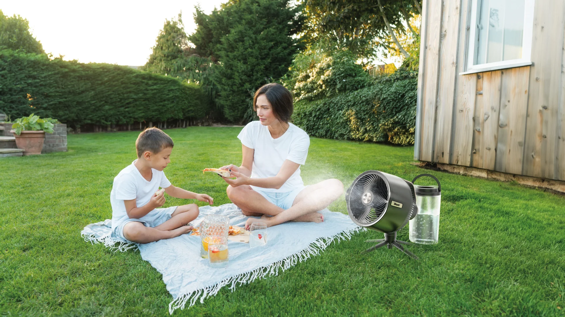 Egy nő és egy gyermek piknikeznek egy takarón az udvaron. Egy ventilátor és egy vizes palack a füvön. Pizza és gyümölcslé a takarón.