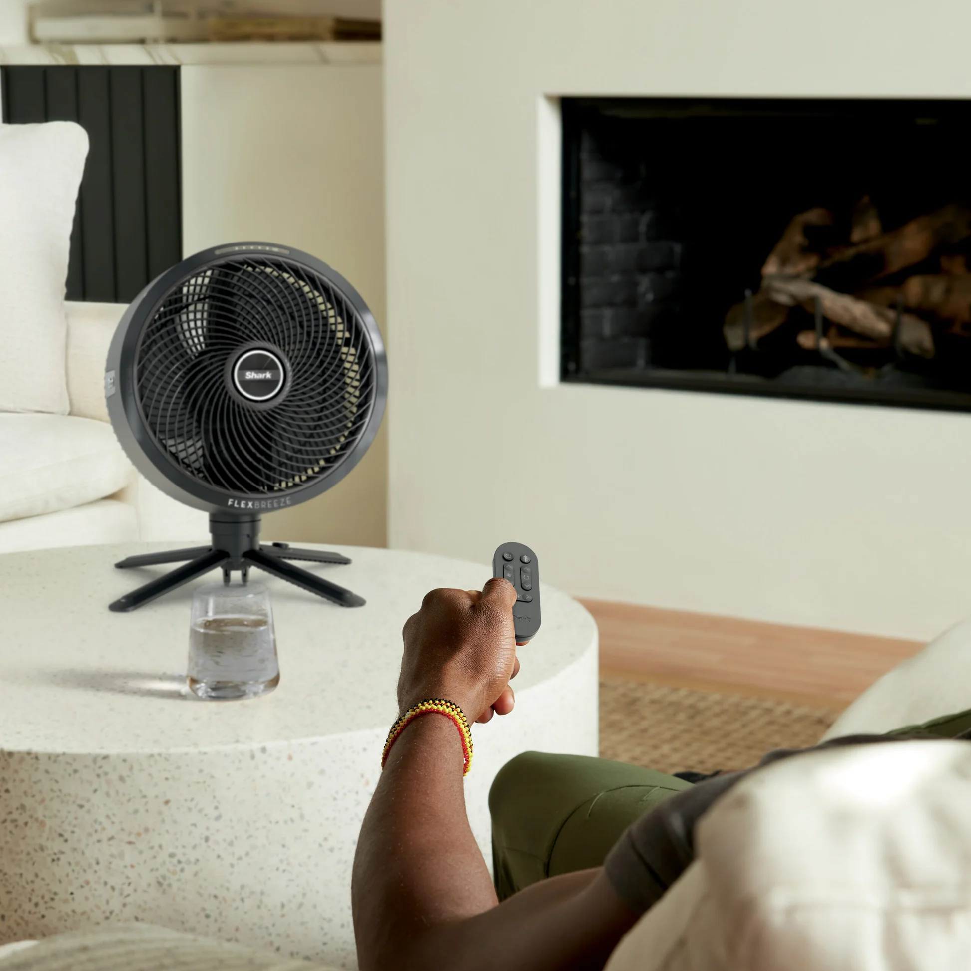 Egy karkötős személy távirányítóval vezérel egy ventilátort. Egy pohár víz és egy kandalló a háttérben.