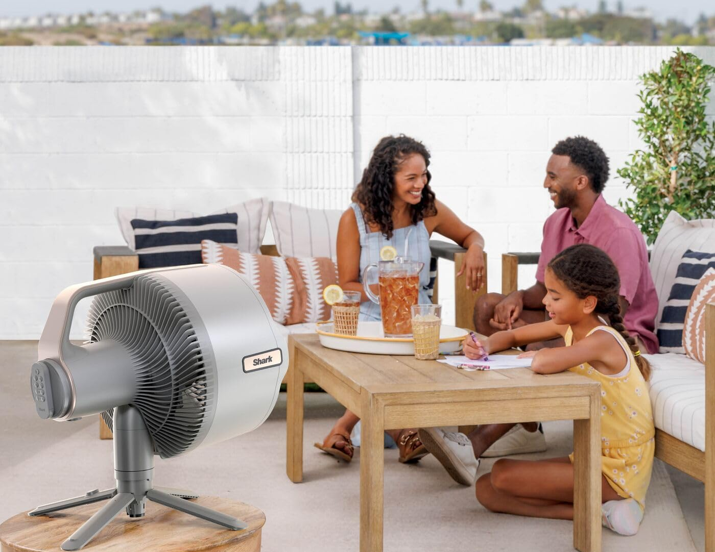 Család a teraszon ventilátorral. Nő, férfi és gyermek egy asztal körül ülnek. Ventilátor előtérben, italok az asztalon, fehér fal a háttérben.