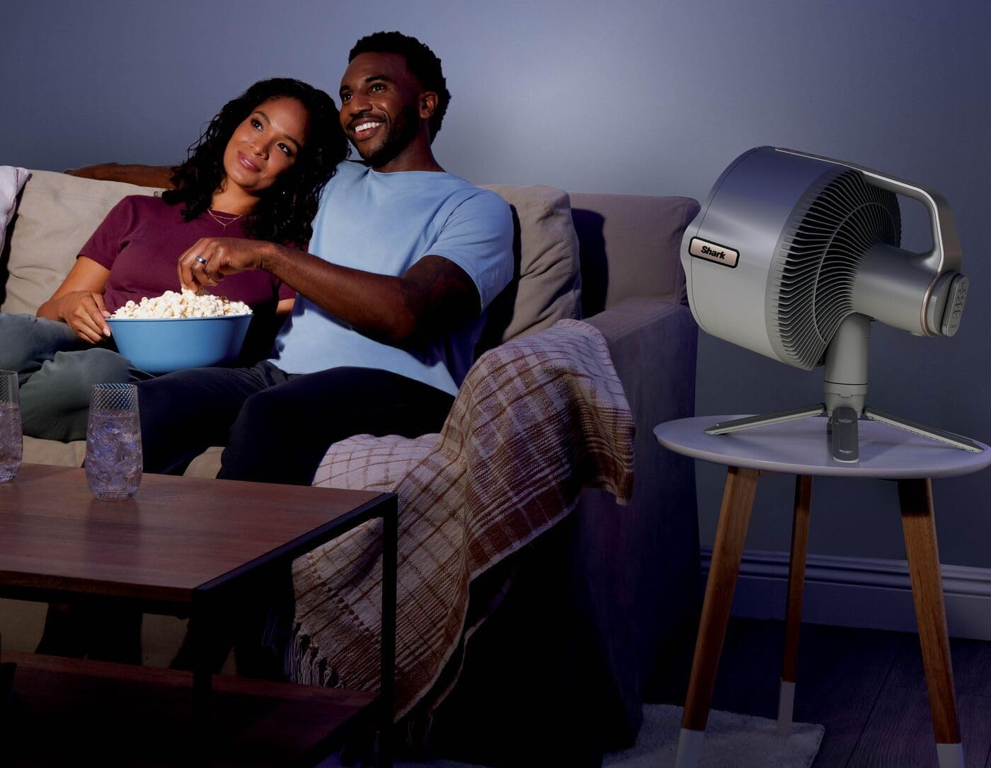 Egy pár a kanapén ülve pattogatott kukoricát eszik. Egy ventilátor egy dohányzóasztalon áll. Egy dohányzóasztal poharakkal az előtérben.