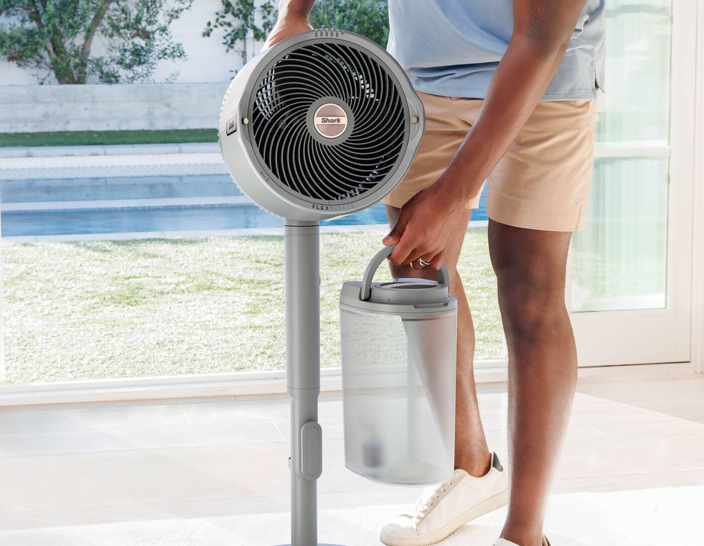 Férfi szürke ventilátort tart víztartállyal. Sortot és kék inget visel. A háttérben medence és kert látható.