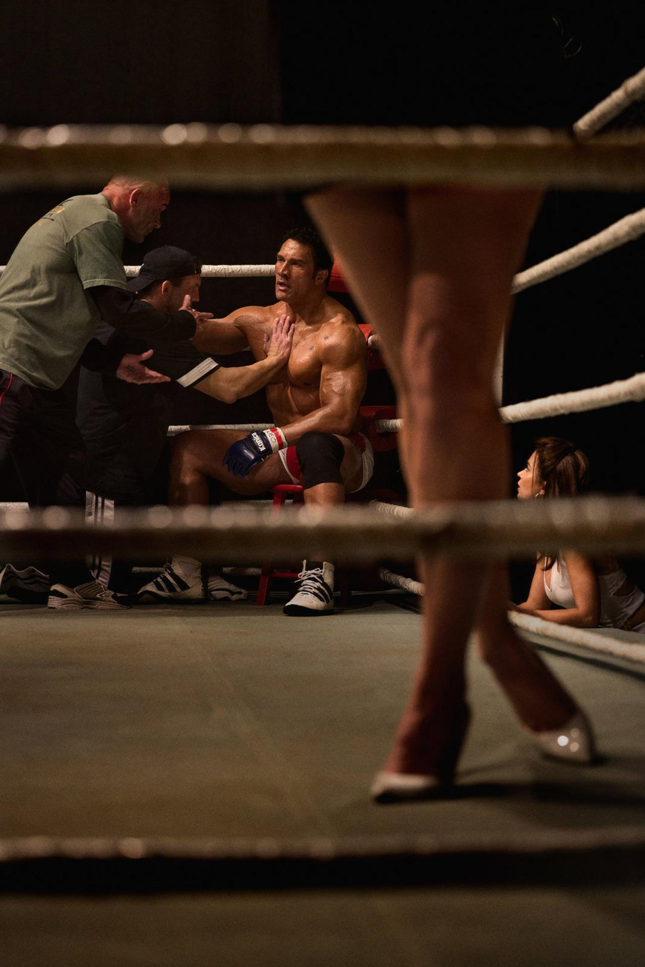 Ein muskulöser Boxer sitzt im Boxring und wird von einem Trainer betreut. Im Vordergrund ist eine Frau mit Absätzen, und eine andere Frau schaut zu.