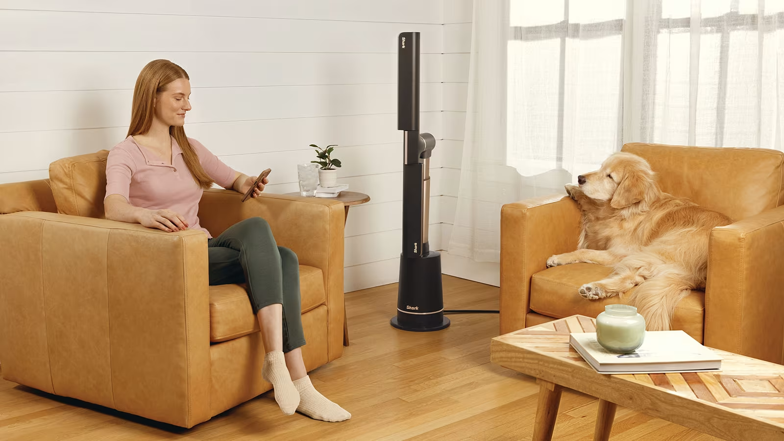 Egy nő egy bézs kanapén ül, és telefont használ. Egy kutya egy másik kanapén pihen. Egy ventilátor áll. Egy asztal van középen.