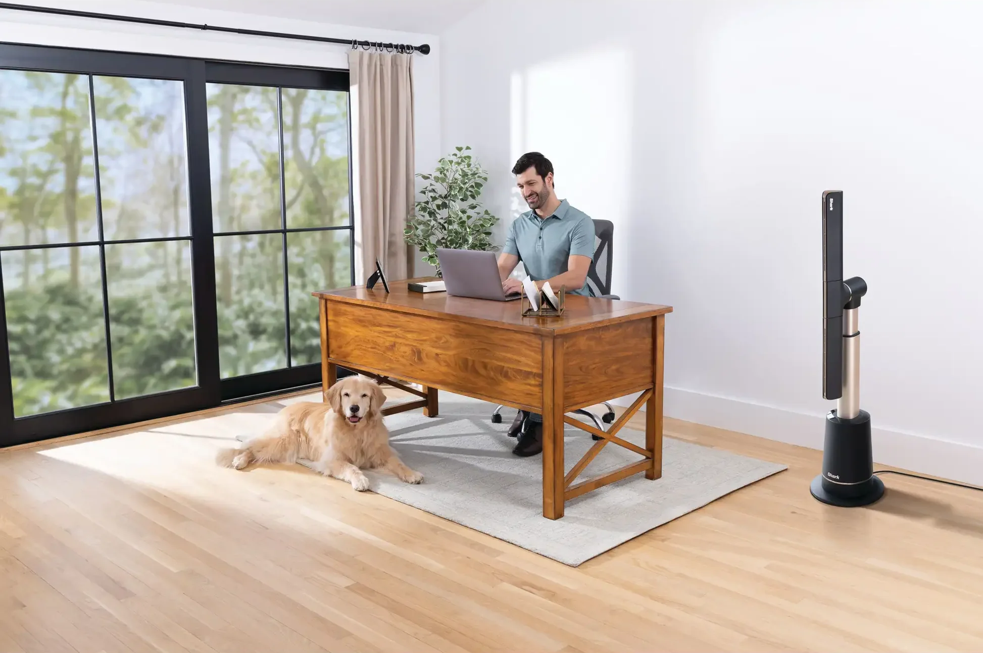 Egy férfi laptopon dolgozik egy faasztalnál. Egy golden retriever fekszik a padlón. Egy ventilátor jobbra. Egy ablak a háttérben.