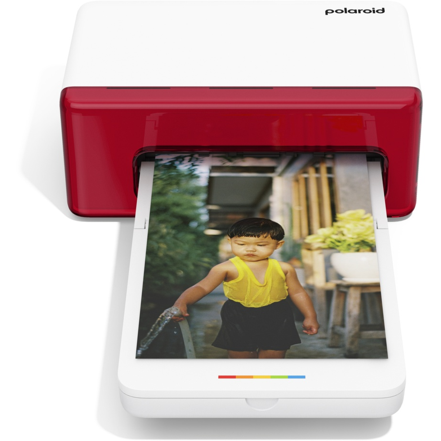 Een Polaroid-printer drukt een foto af van een kind in het geel, met een slang. De achtergrond toont planten, een houten kruk en een huis.