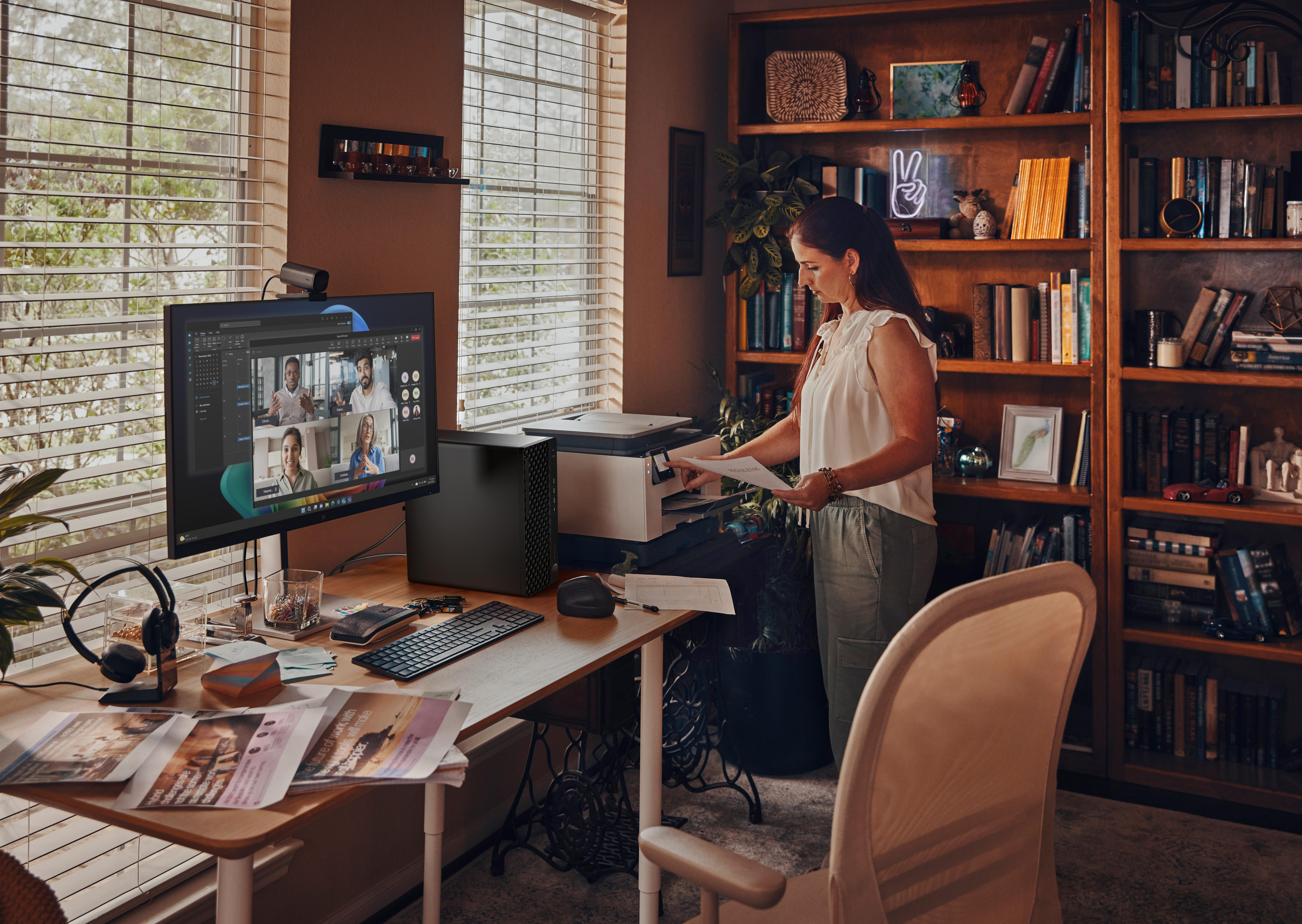 Frau im Homeoffice, betrachtet Papiere in der Nähe eines Druckers. Desktop-Computer auf dem Schreibtisch, Videoanruf sichtbar. Bücherregal im Hintergrund.