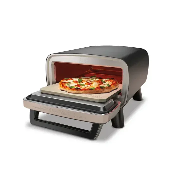 Egy pizza egy fekete pizzasütőben lévő kőre van helyezve. A pizzán sajt és bazsalikom van. A sütő fehér háttér előtt van.