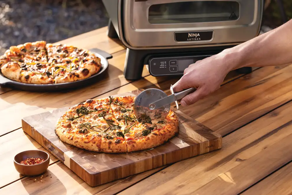 Egy pizzát egy fa deszkán vágnak. Egy másik pizza a háttérben van. Egy pizzakemence látható. Egy kis tál chili pehellyel.