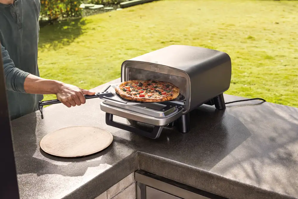 Egy személy egy fekete pizzasütőből vesz ki pizzát. A háttérben egy pizzakő és egy zöld udvar látható.