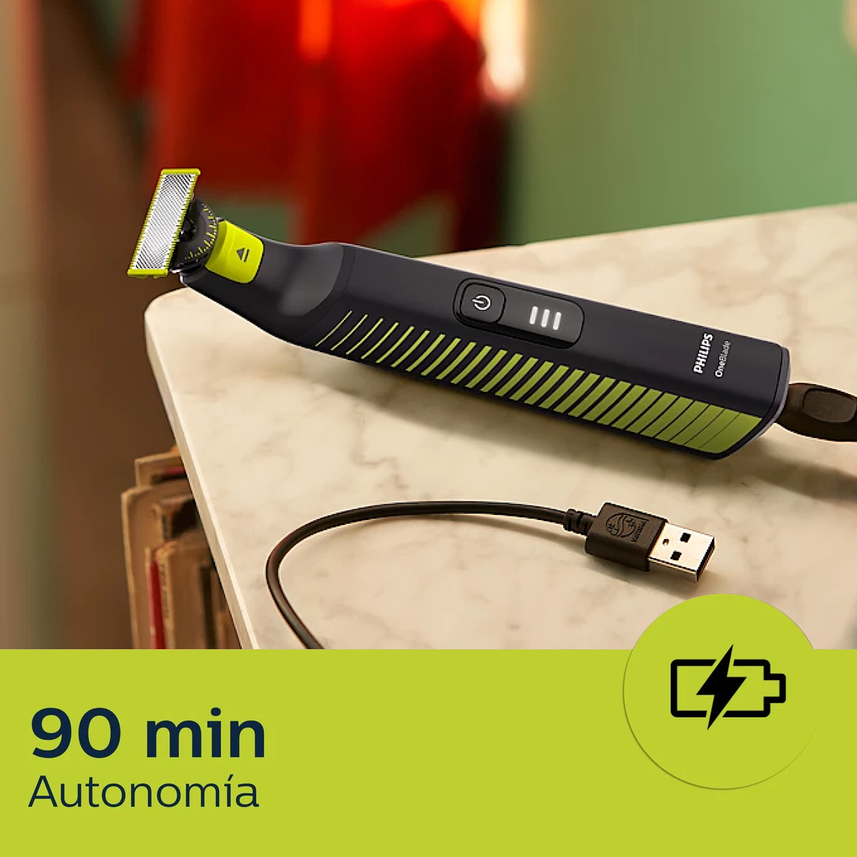 Recortadora Philips OneBlade negra y verde con cable USB sobre una superficie de mármol, junto a un libro. Icono de batería y '90 min Autonomía'.