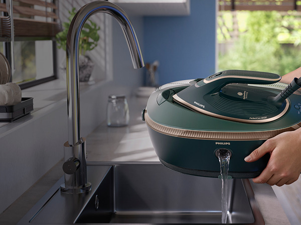 Una persona vaciando una plancha Philips PerfectCare 8000 verde azulado en un fregadero. Un grifo plateado es visible a la izquierda. Plantas verdes al fondo.