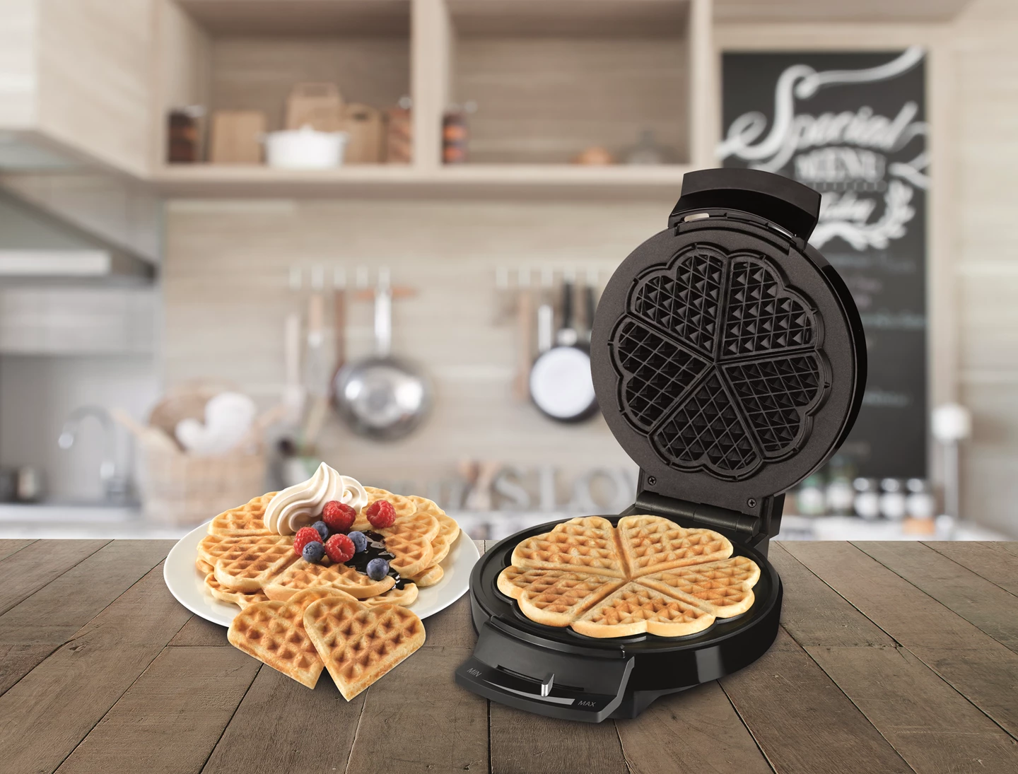 Una macchina per waffle con un waffle cotto. Un piatto di waffle con frutti di bosco e panna. Lo sfondo è una cucina.