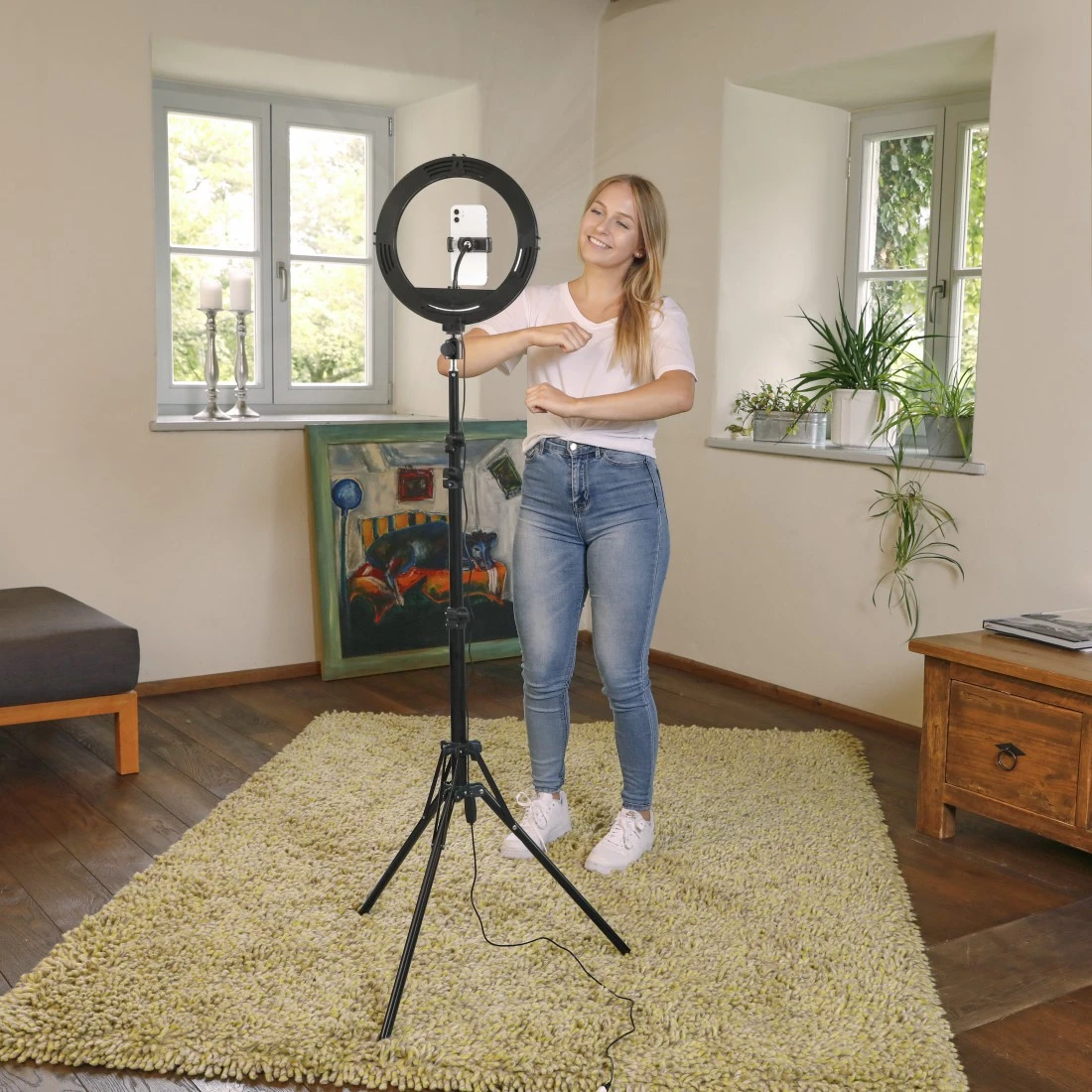 Eine Frau steht neben einem Ringlicht und filmt mit einem Telefon. Sie trägt Jeans und ein weißes Hemd und lächelt. Teppich, Fenster und Kunst sind sichtbar.