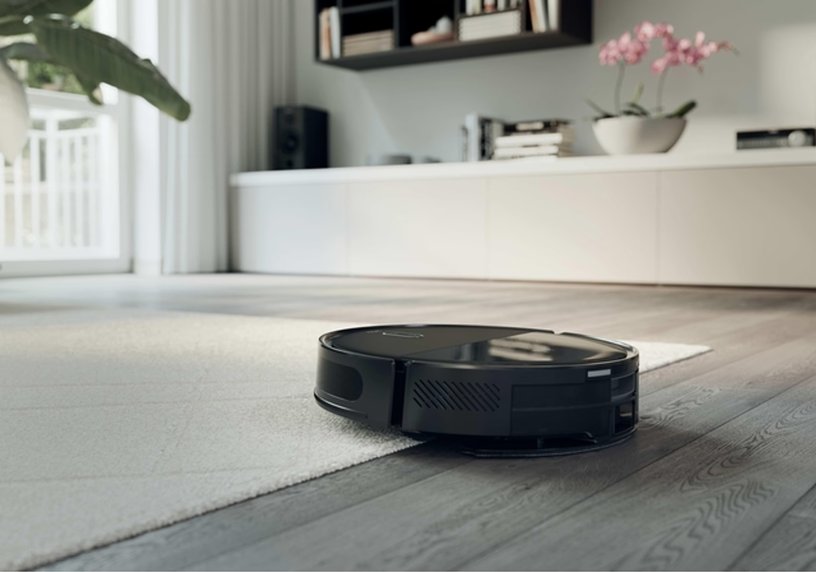 Un aspirateur robot noir sur un tapis dans un salon moderne. L'arrière-plan comprend une fenêtre, des étagères et une plante.