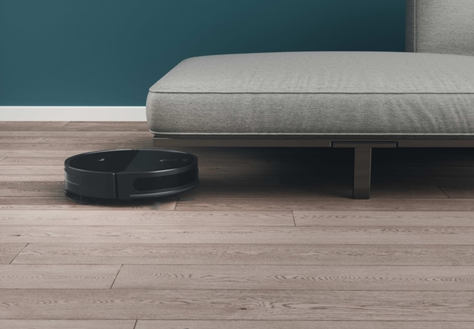 Un aspirateur robot noir sur un sol en bois. Un canapé gris est en arrière-plan contre un mur bleu-vert.