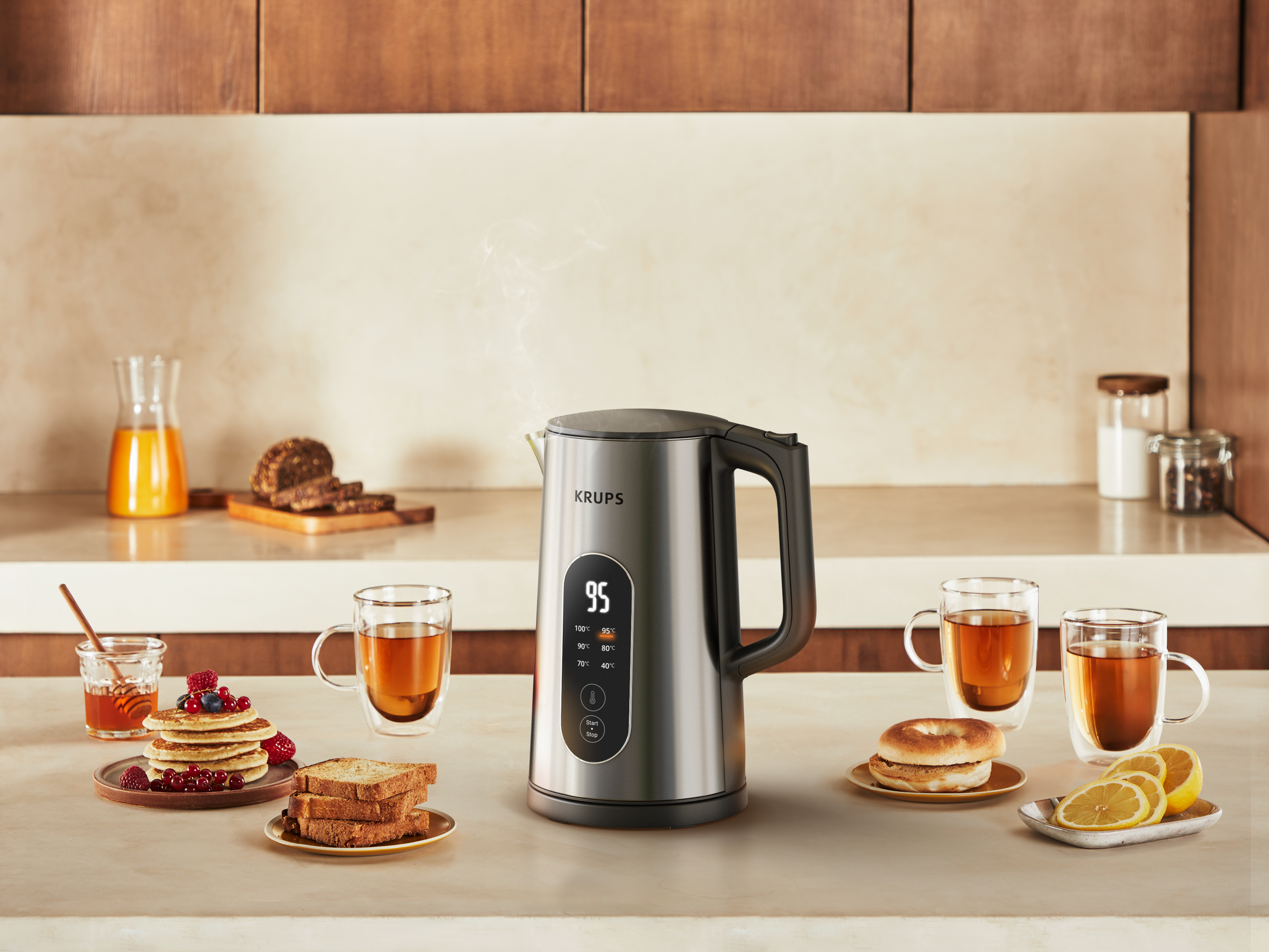 Eine Küchenszene mit einem Wasserkocher, Essen und Getränken. Pfannkuchen, Toast, Bagels, Zitronen und Teetassen sind sichtbar.