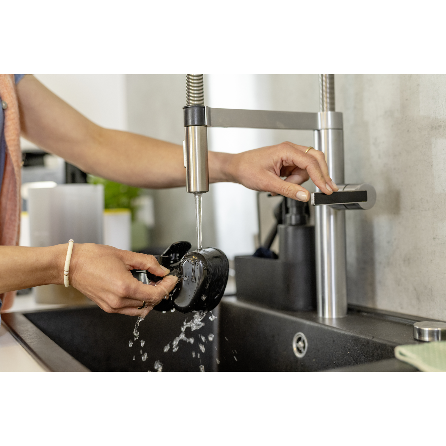 A person washes a black coffee maker under a running faucet in a kitchen sink. Water streams down, creating splashes.