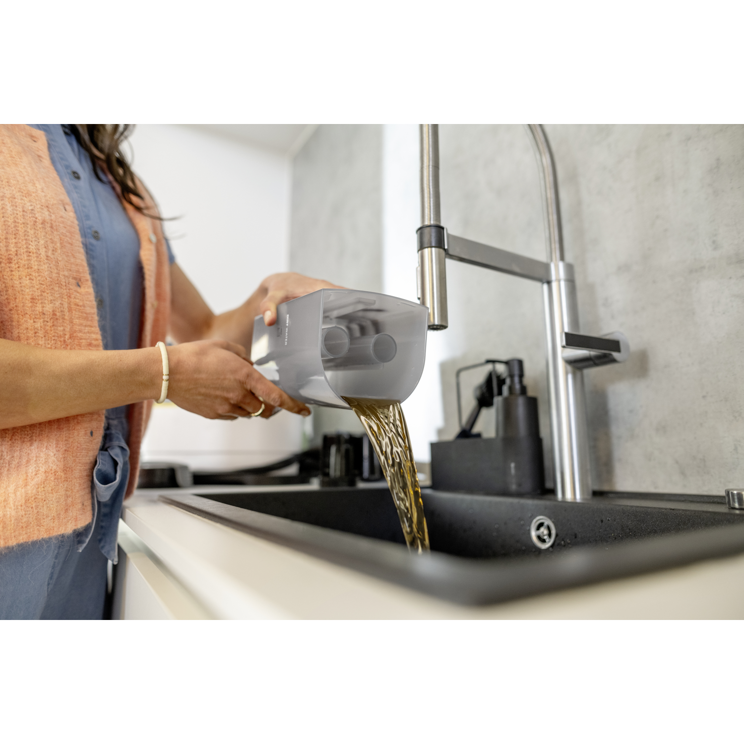 A person pours liquid from a gray container into a dark sink in a kitchen. A faucet is in the background. The person wears a blue shirt and orange sweater.