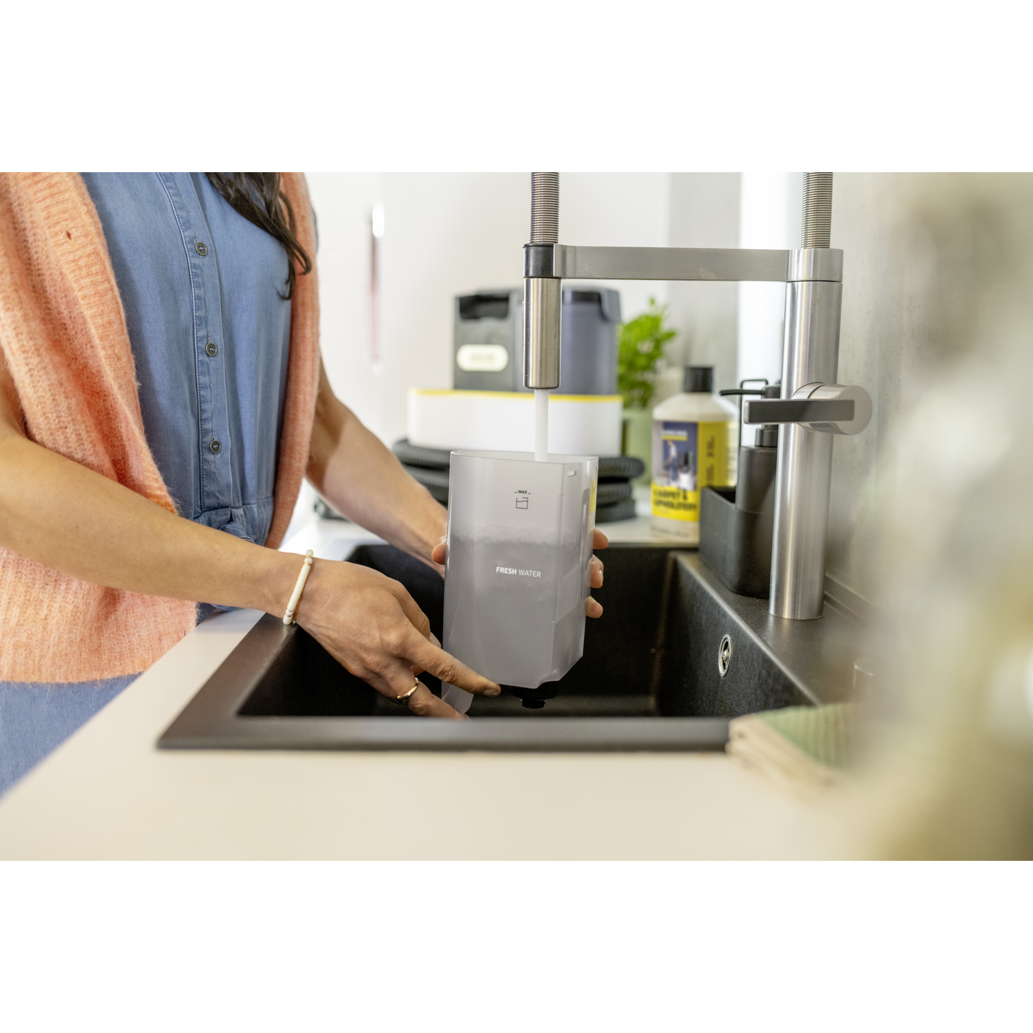 A woman fills a water container under a faucet. The container is labeled 'Fresh Water'. Sink is black, and woman wears a bracelet.